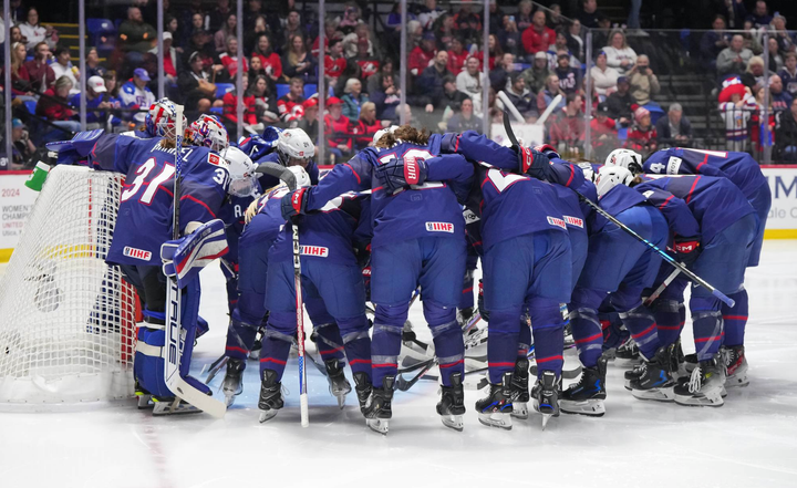 Team USA hunches over, arm-in-arm, in a huddle in front of their goal before the gold medal game. They are wearing blue.