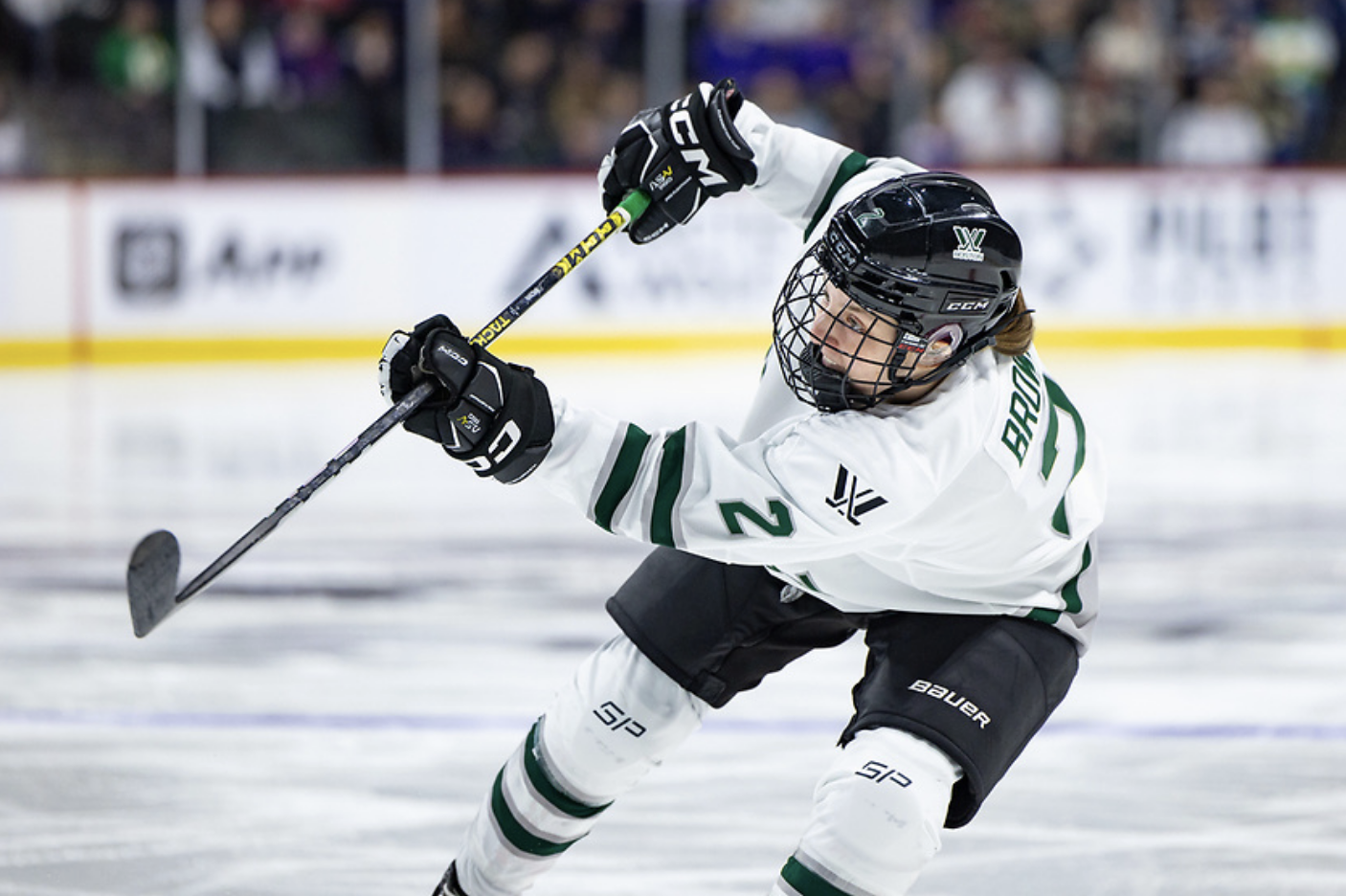 Brown leans into a shot follow through. She is wearing a white away jersey and patrolling the left side of the blue line.