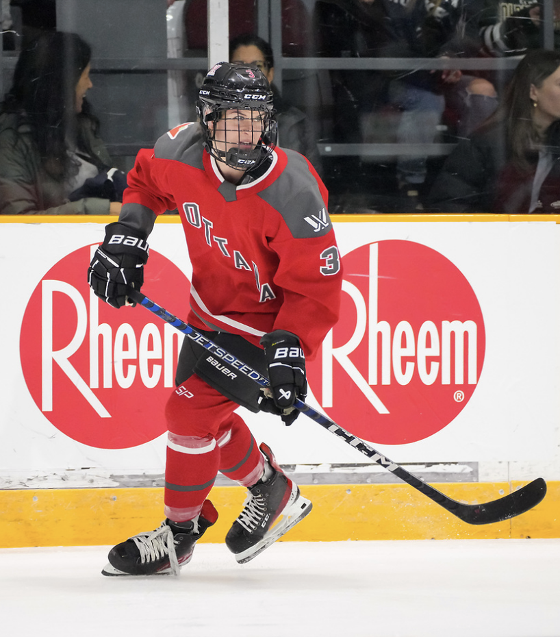 Boyd skates with both hands on her stick and watches the play to her left. She is wearing a red home jersey.