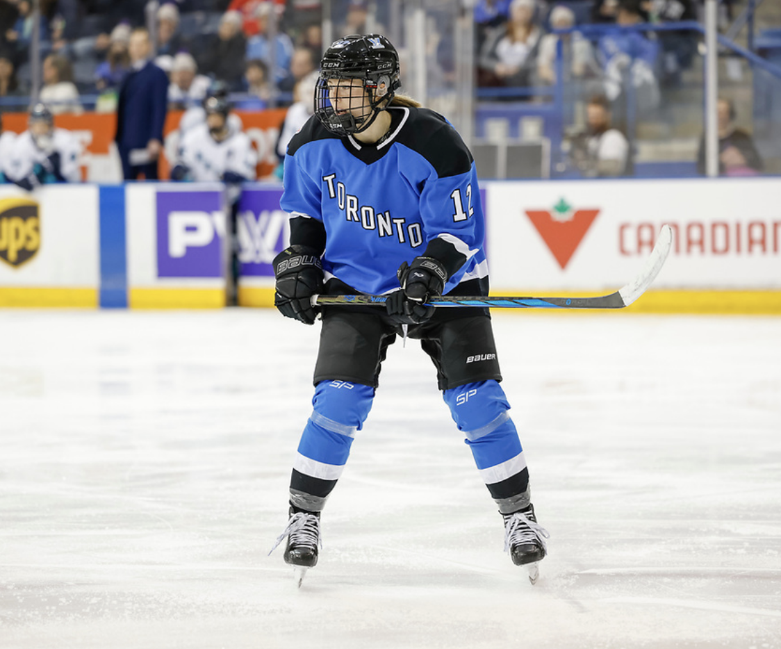 Munroe watches a play unfold during a game. She is holding her stick at waist level and looking to her right while wearing a blue home uniform.