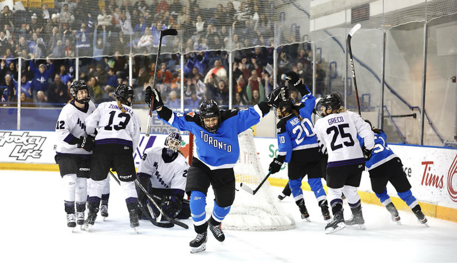 Spooner celebrates raises her arms and celebrates a goal as she skates away from the net. She is wearing a blue home uniform.