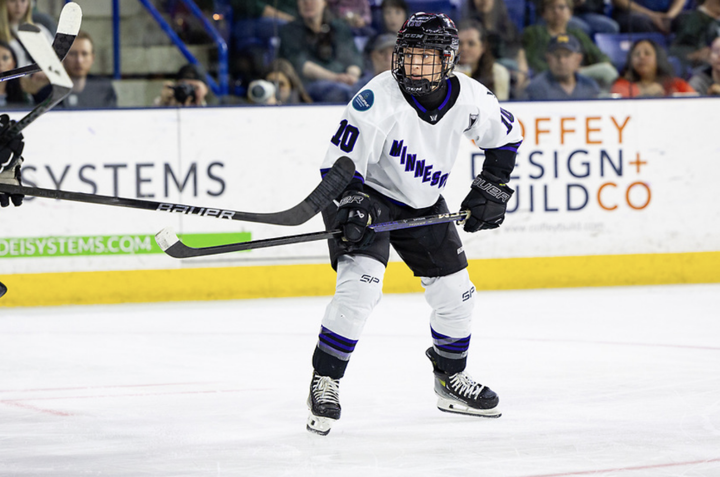 Brodt dishes the puck. Her stick is at her waist and she is looking to the right. She is wearing a white away uniform.