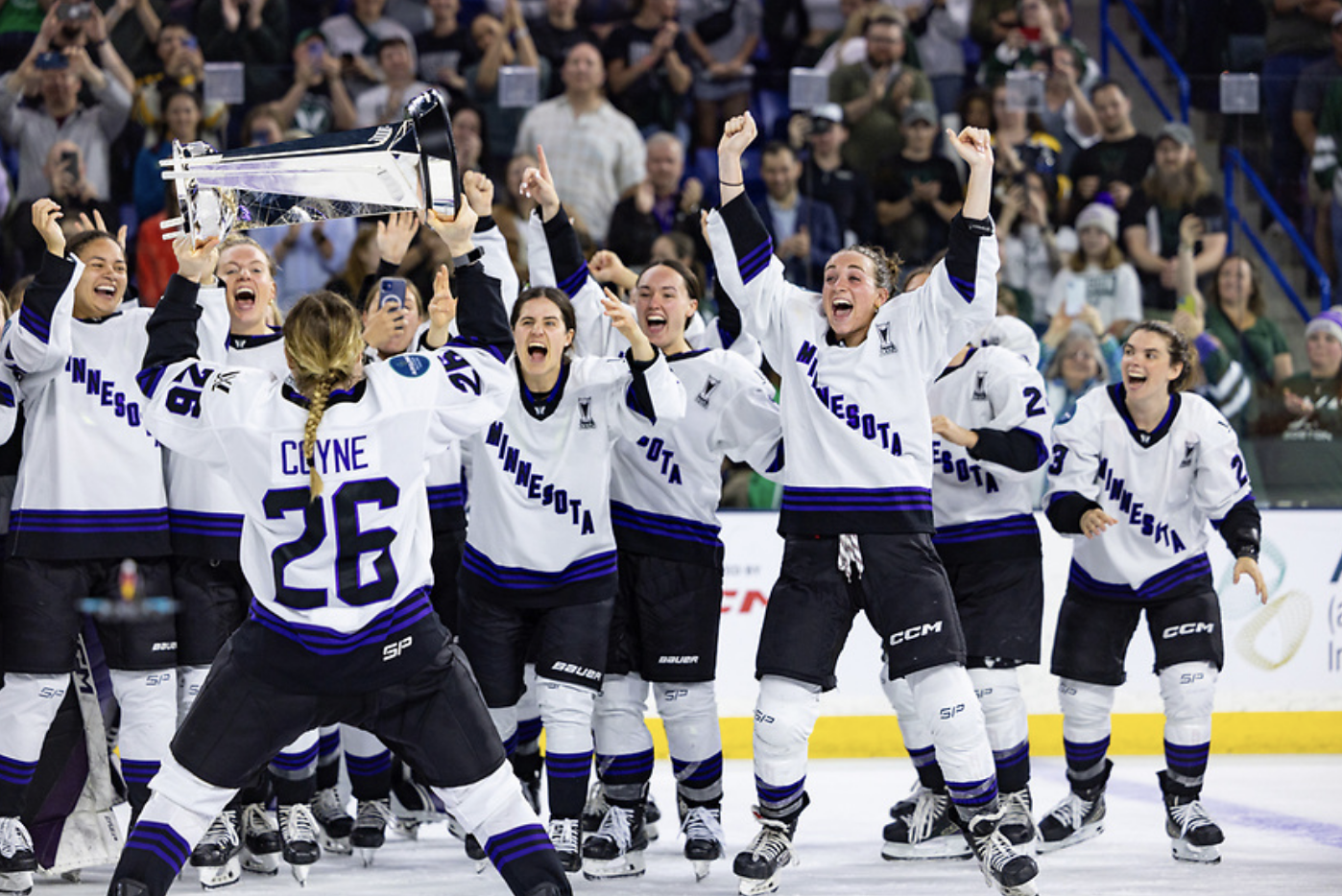 Coyne Schofield raises the Walter Cup as she skates to her teammates, who are cheering. Most of them also have their arms raised. They are all wearing white away uniforms.