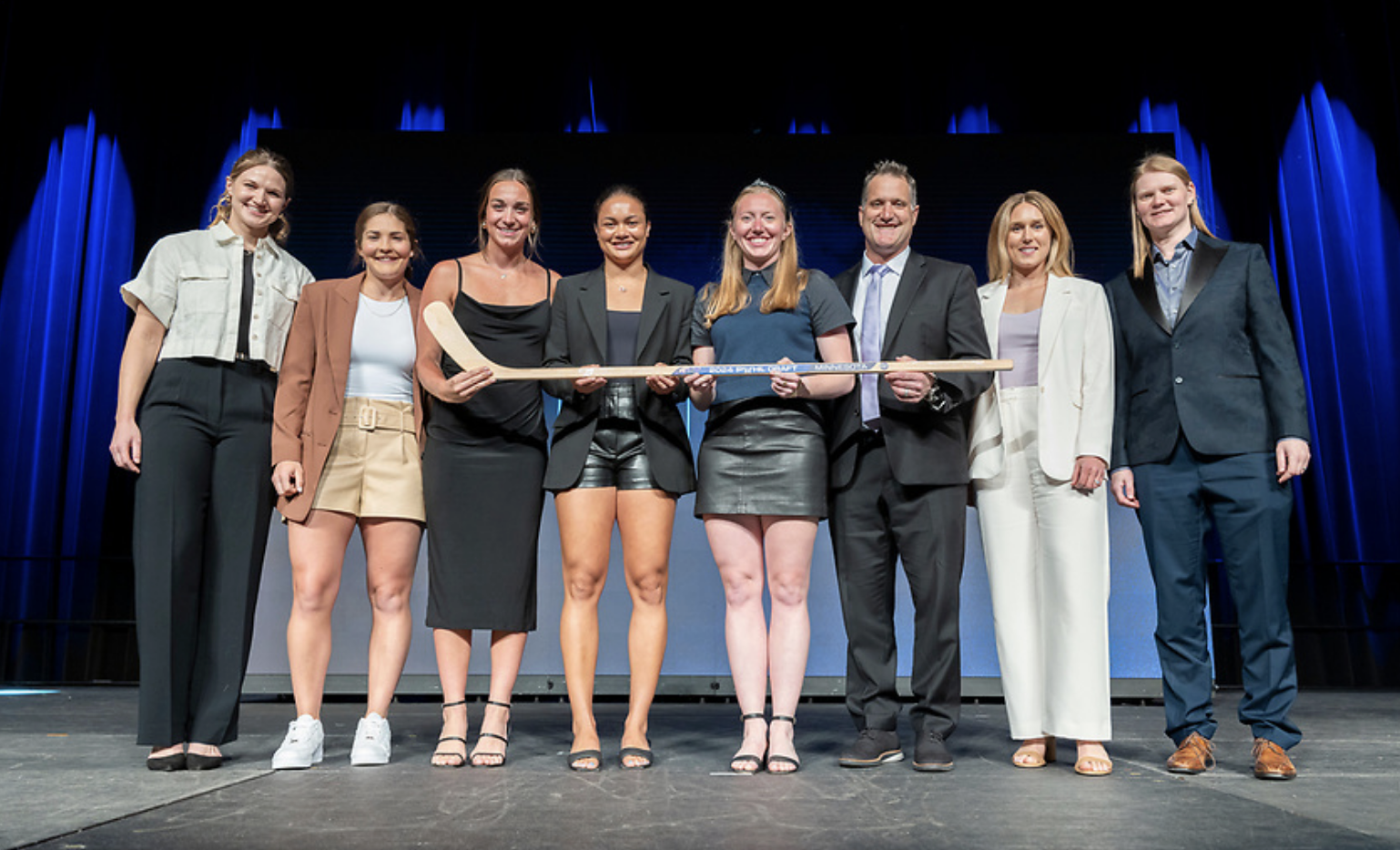 Eight representatives of PWHL Minnesota pose for a photo on stage while holding a wooden stick at the 2024 draft. 