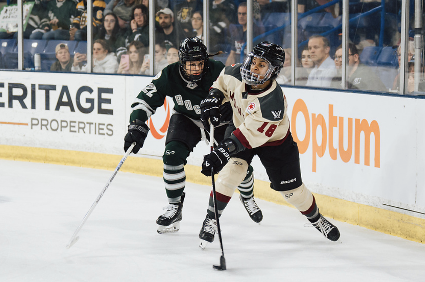 Grant-Mentis extends her arm to pass the puck from behind the net while wearing a cream away jersey. A Boston defender is reaching around her.