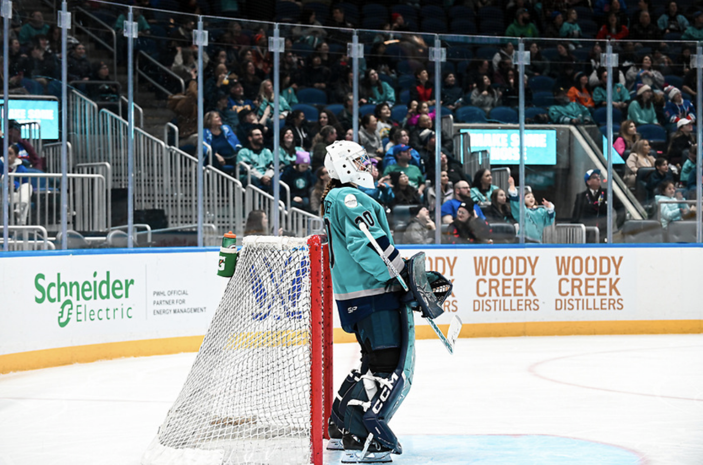 Schroeder stands in front of her net and looks up ice. She is wearing a white mask and teal home uniform.