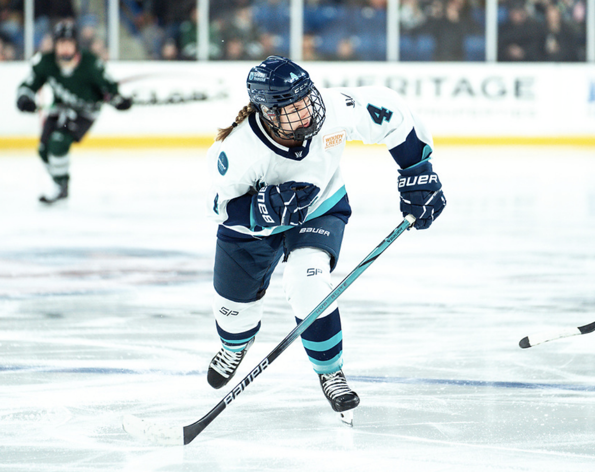 Baker skates leaning forward. She is looking to the left and wearing a white away uniform.