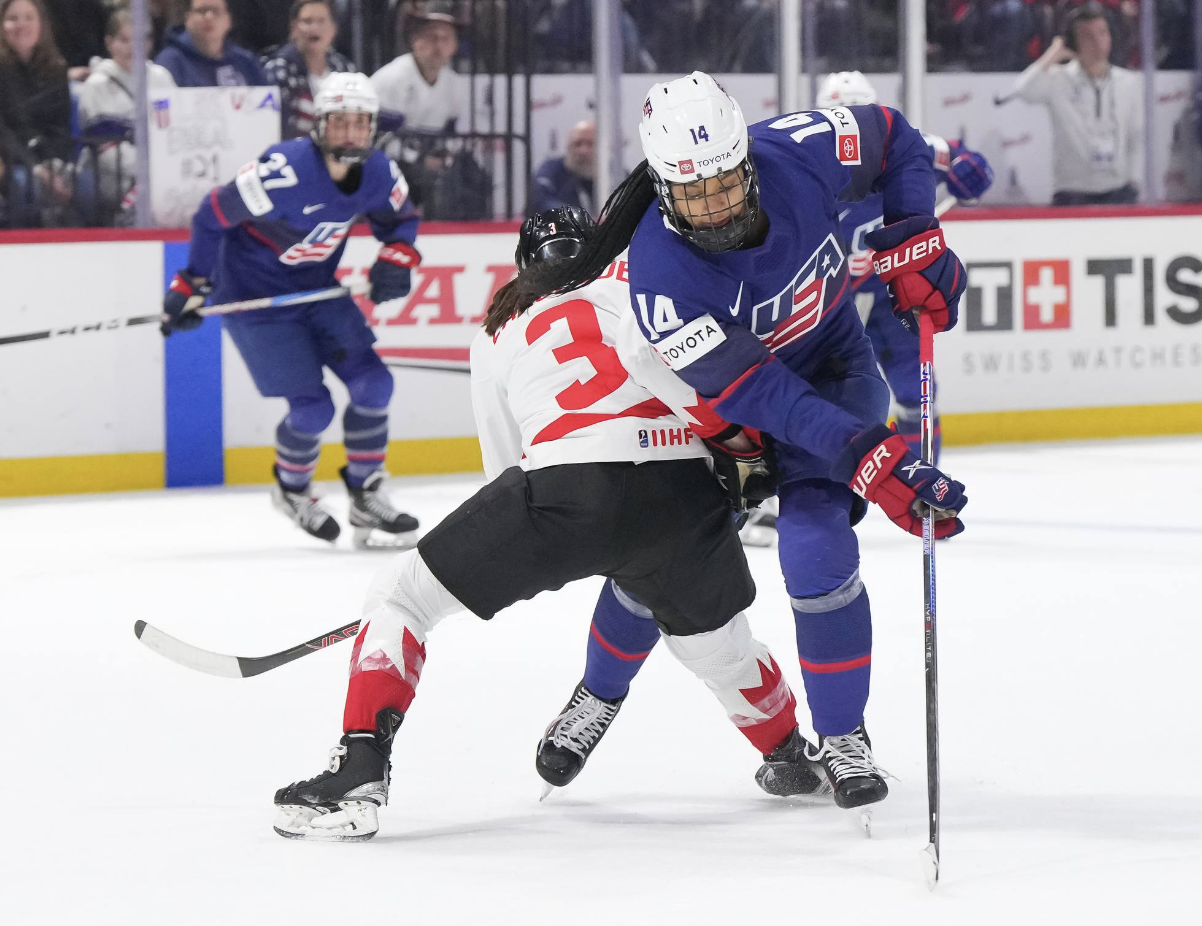 Edwards protects the puck as she attempts to skate around Larocque, who has made contact with her. Edwards is wearing blue and skating towards the camera, while Larocque is wearing white and back to the camera.