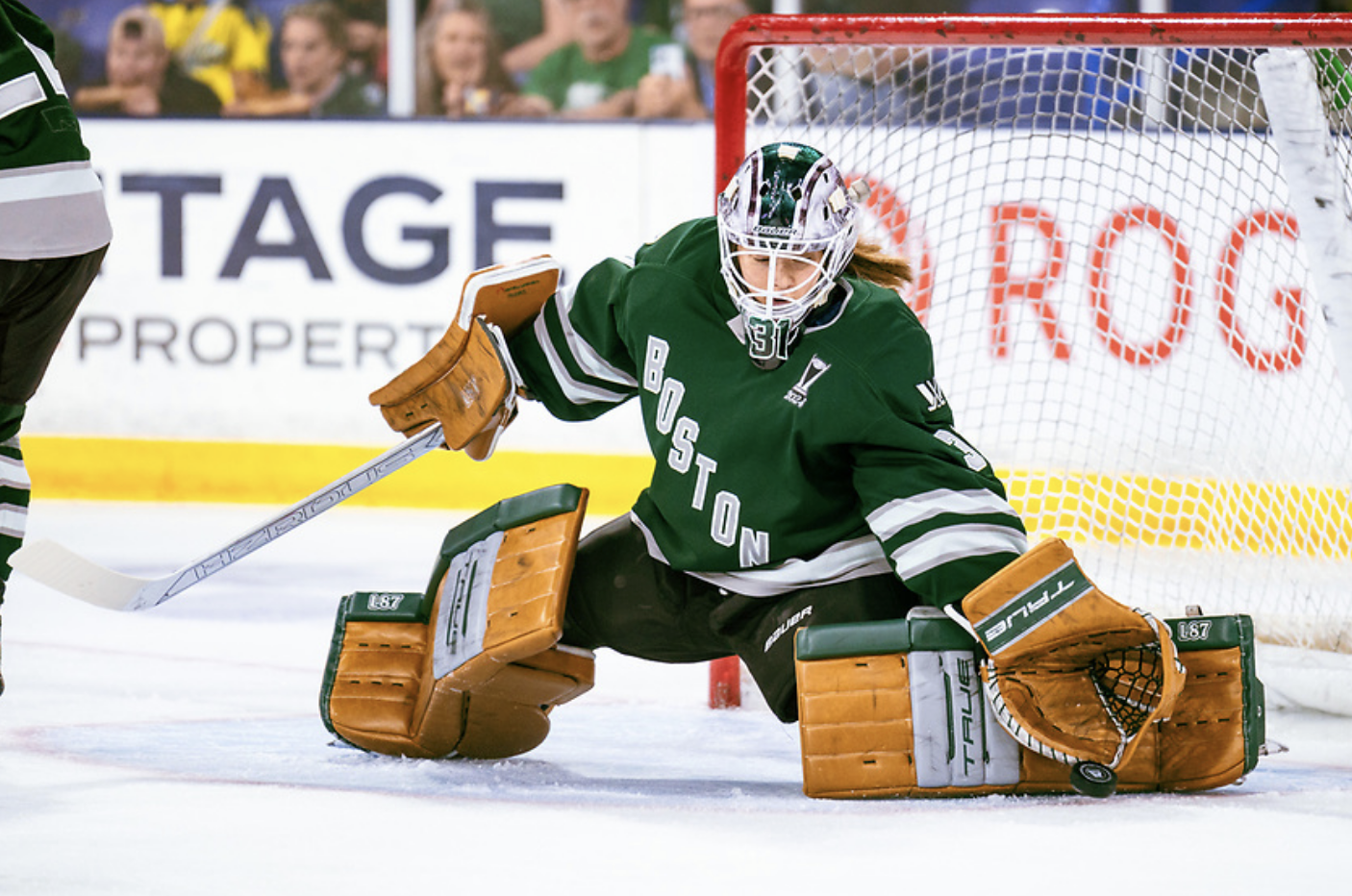 Frankel pushes to her left from her knees and reaches down with her glove to make a save. She is wearing light brown and green pads, a green and white mask, and a green home jersey.