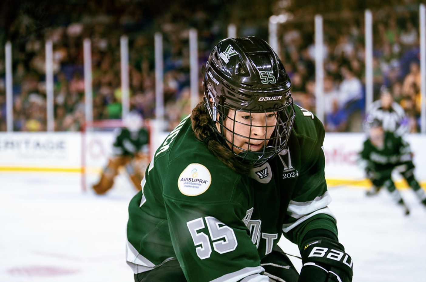 Babstock looks down at the out-of-frame puck, trying to scoop it out from along the boards. She is skating up ice wearing a green home jersey. 