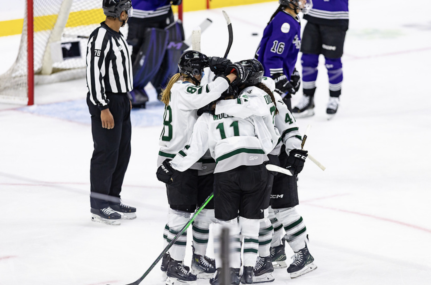 Five Boston players celebrate a goal with a tight group hug in the left faceoff circle. They are all wearing white away jerseys.