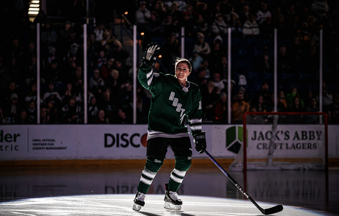 Marvin is standing in the spotlight and looking up at the crowd, right arm raised in a wave. She is wearing a green home uniform.