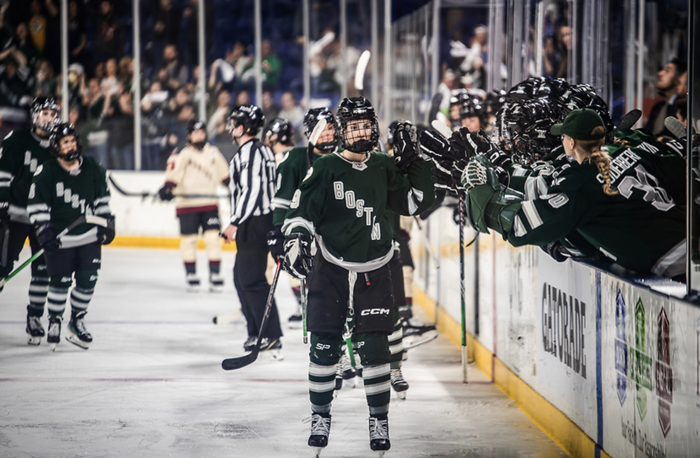 Shirley faces the camera and leads the handshake line at that bench as players lean over for fistbumps. They are all wearing green home uniforms.