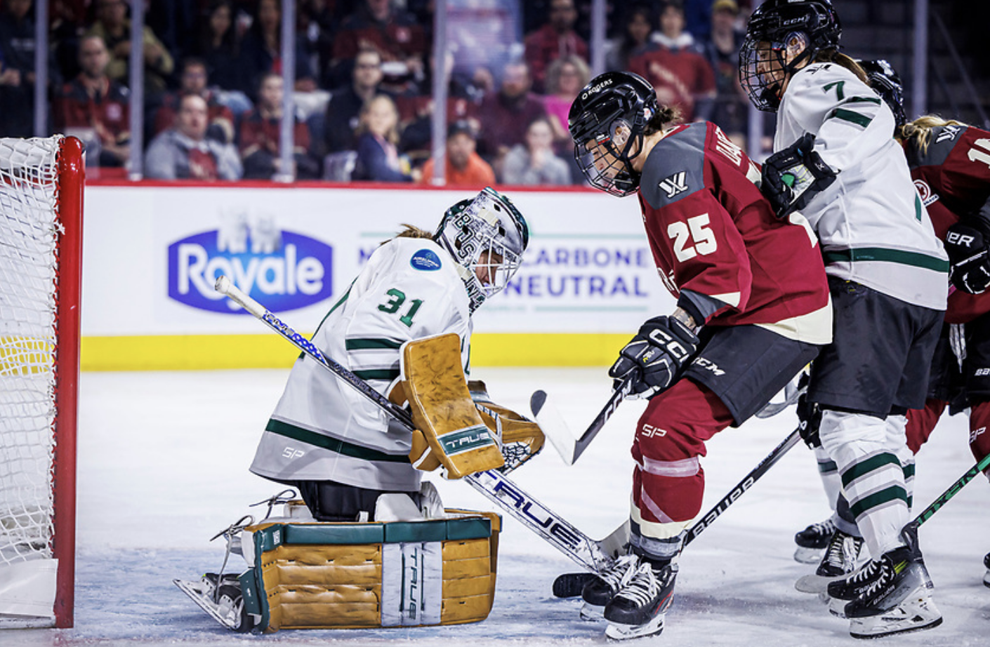 Frankel is on her knees, leaning slightly forward to make a save. Montréal's Mélodie Daoust is standing right in front of her, while Sidney Morin pushes her from behind. The Boston players are wearing white, while Daoust is wearing maroon.