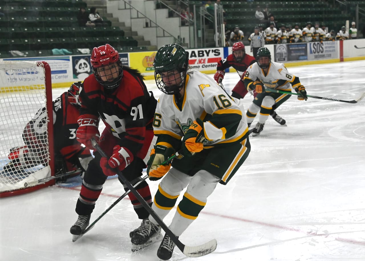 This photo is taken from behind the end zone. The Oswego players are wearing their home whites, while Plattsburgh is dressed in red with black trim. In the center of the photo, one player from each team is headed towards the end boards, their sticks crossed, while another player from each team skates towards the action. The Plattsburgh goalie is crouched in her net on the left edge of the photo.