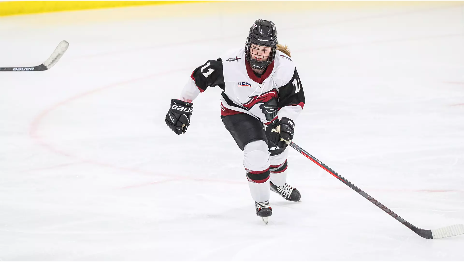 This player is right-handed, and holds her stick at the top with her left hand. Her helmet, gloves, and pants are black. Her jersey, #11, is white with black bands at the tops of the sleeves, and a red and black crest on the front. Her white socks have black and red stripes mid-shin.