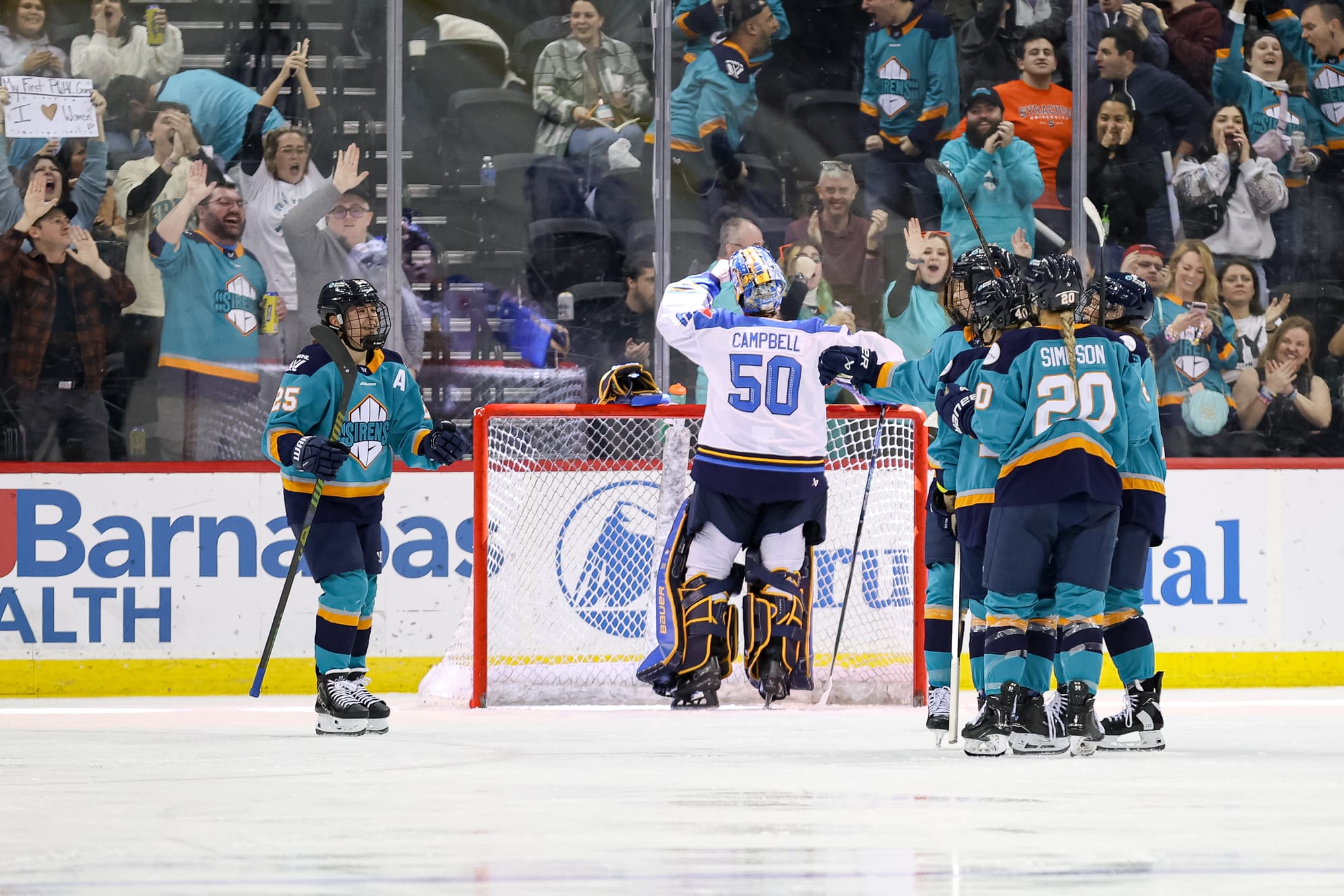 Four players are off to the right in a group hug. Another is skating in to join. They are wearing teal home uniforms. Kristen Campbell is in white standing in the net with her back turned, about to get a drink of water.