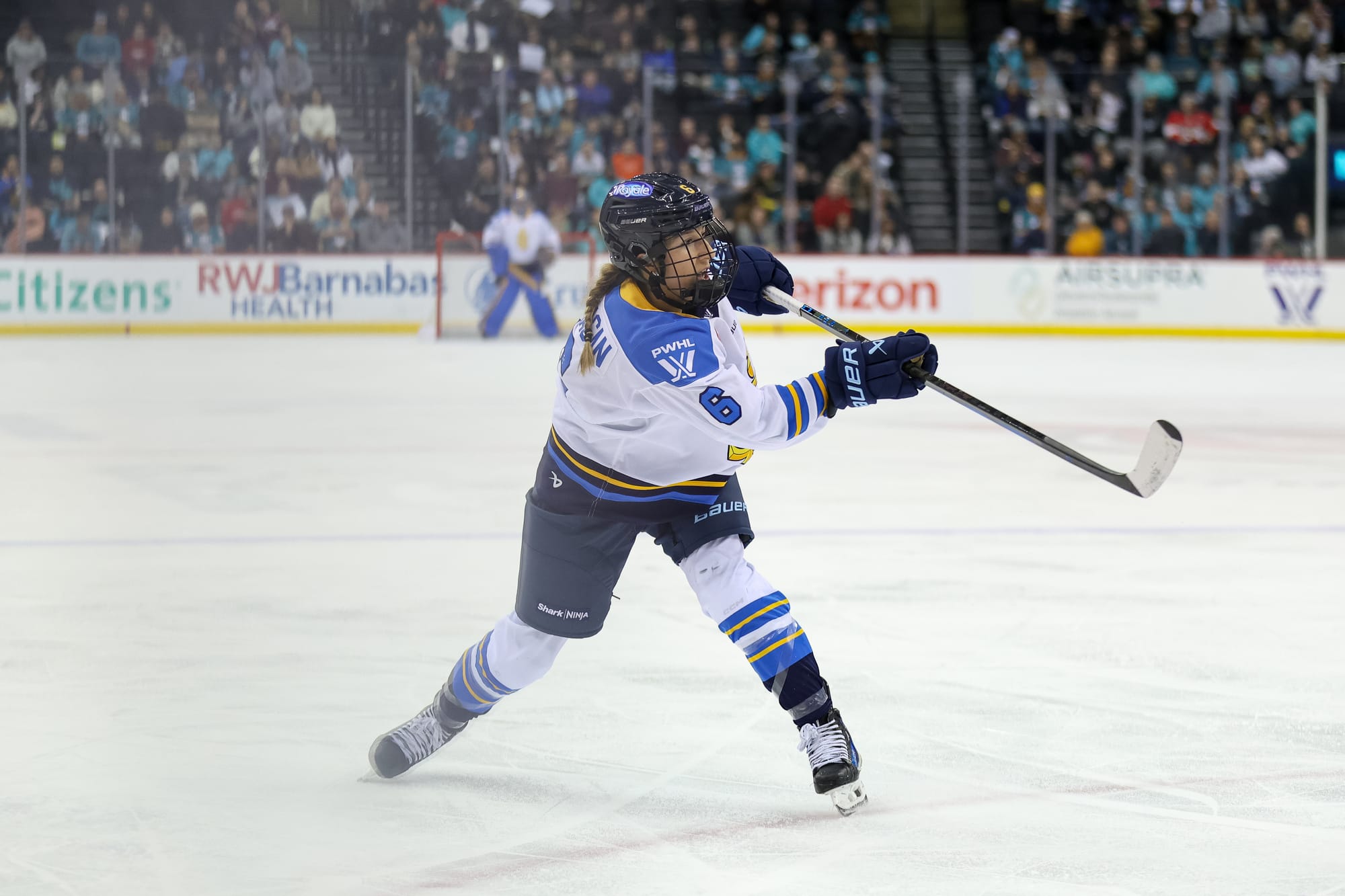 Flanagan is shifting towards her left leg, raising her stick to follow through on a shot. She is wearing a white away uniform.