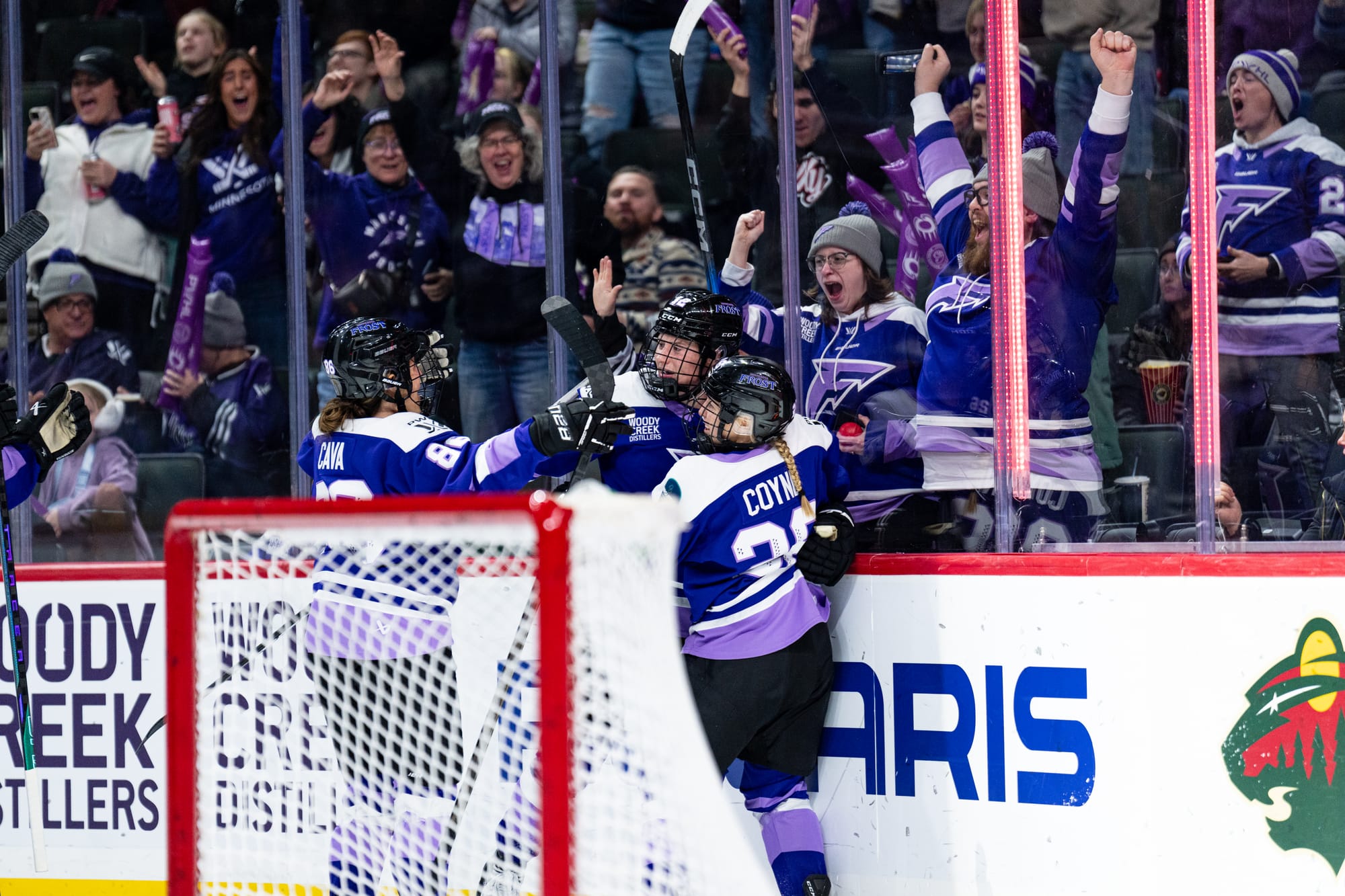 Three Minnesota players celebrate with a group hug behind the goal. They are wearing purple home uniforms. Fans are also celebrating in the background.