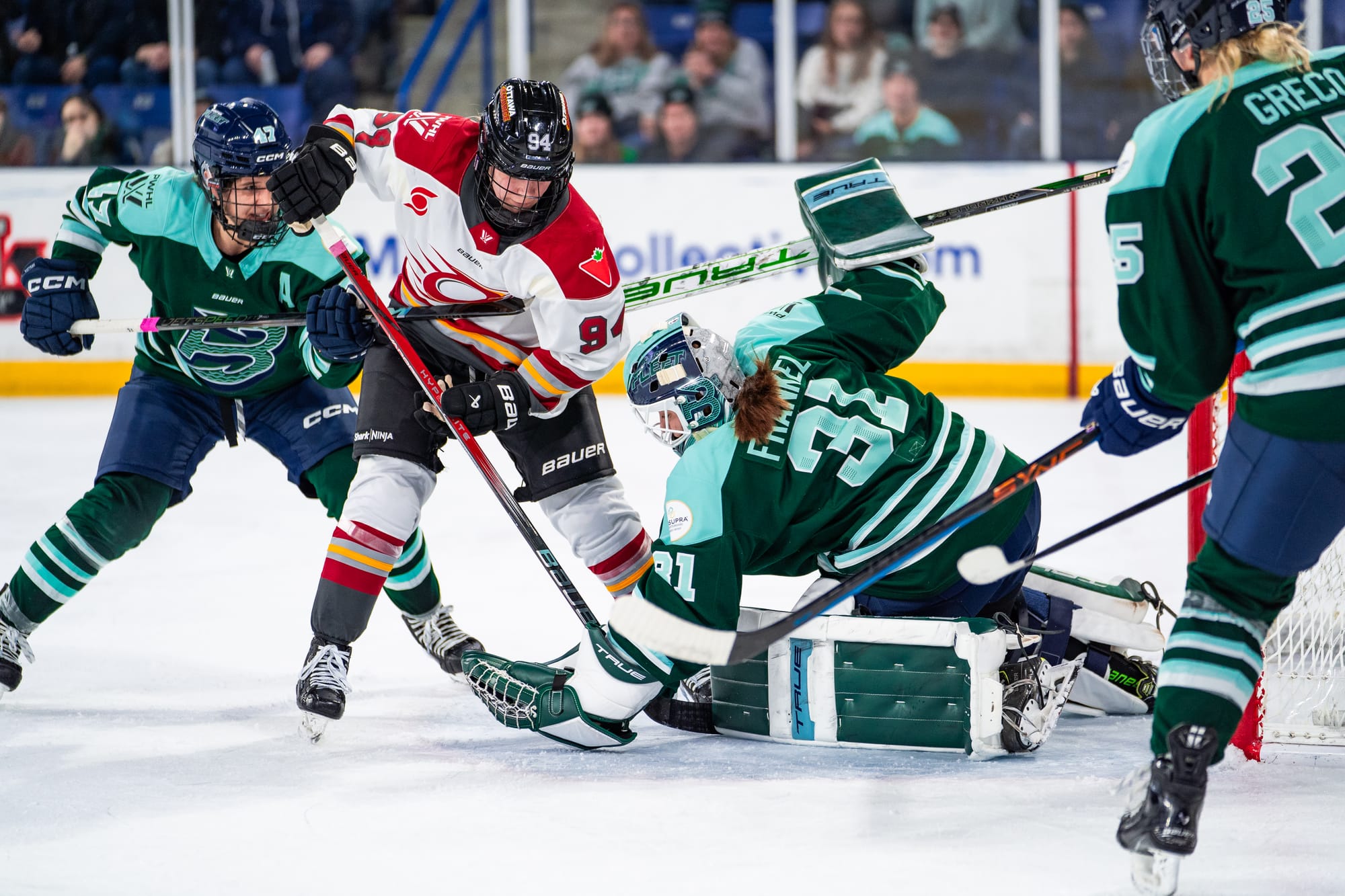 Meixner stands just outside the crease, with Jamie Lee Rattray (who is behind her) and Aerin Frankel's sticks holding her back. From her knees, Frankel reaches forward and to the left to make a save. Meixner is wearing white, while Rattray and Frankel are in green.