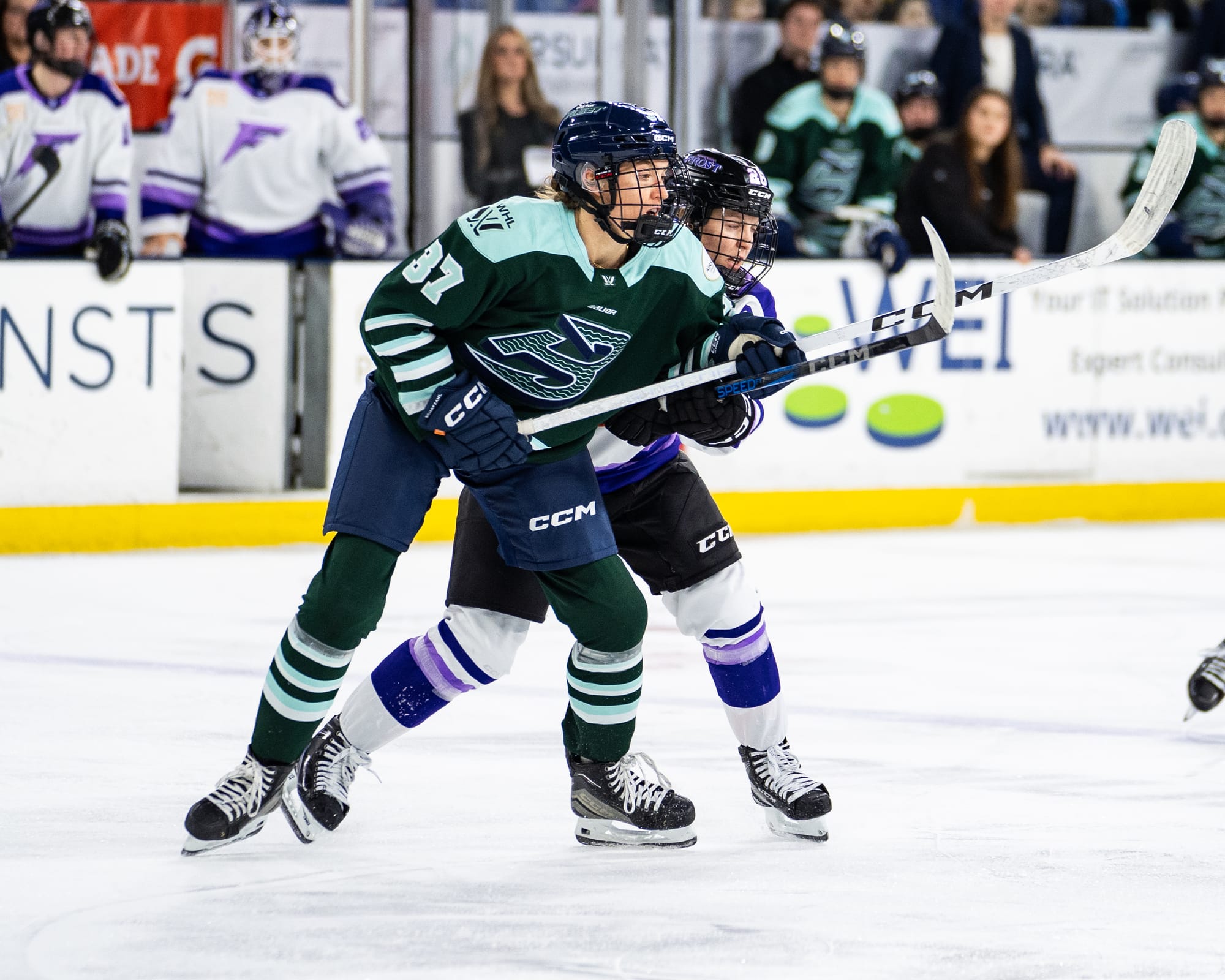 Coyne Schofield makes contact with Shirley from behind, as they battle for positioning. Both have their sticks raised above their waist. Shirley is in a green home uniform, while Coyne Schofield is in white.