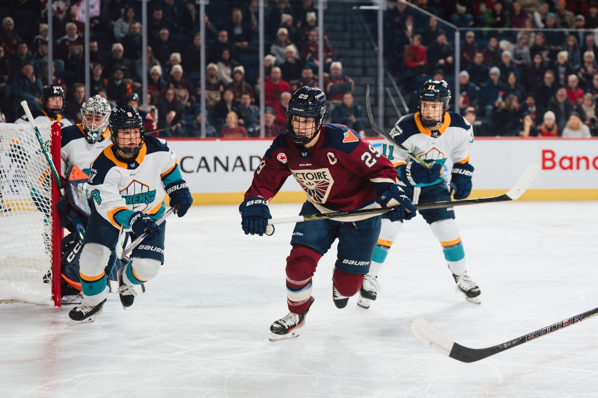 Poulin is looking to her right and skates toward the camera in pursuit of the puck. She is wearing a maroon home uniform. A New York player is chasing her.