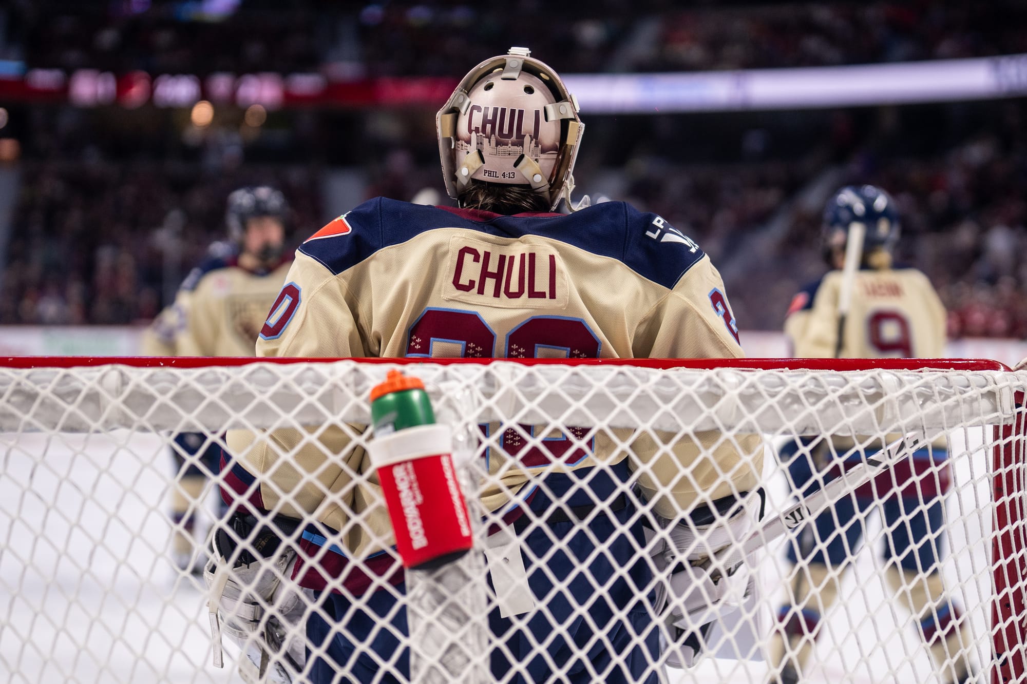 Chuli stands in the middle of the net with her back turned to the camera. Other players are out of focus in the background. They are all wearing cream away uniforms.