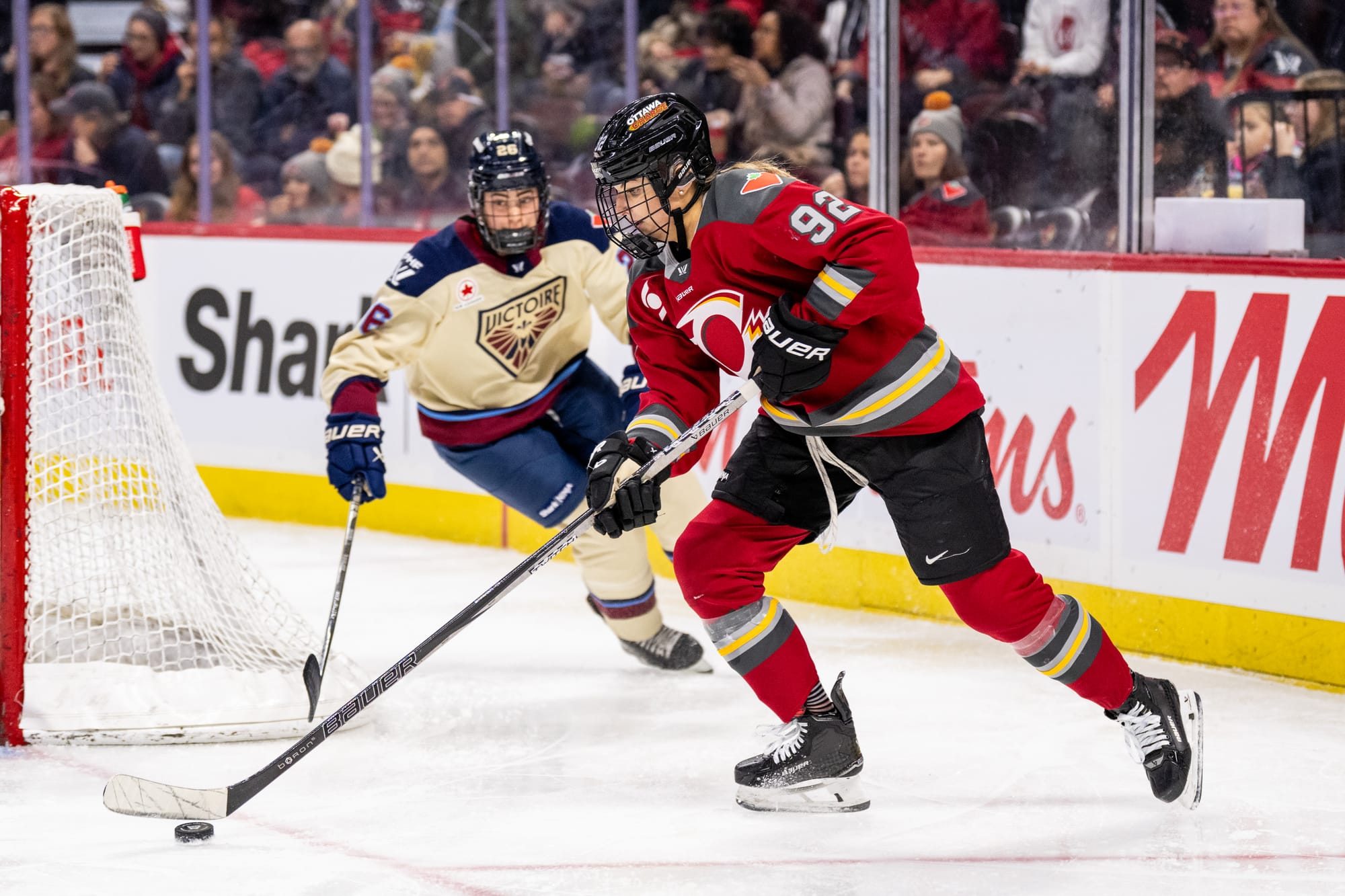 Serdachny (right in red) looks down at the puck as she skates out from just inside the trapezoid. A Montréal player (left in cream) is coming in to try to poke poke the puck away.