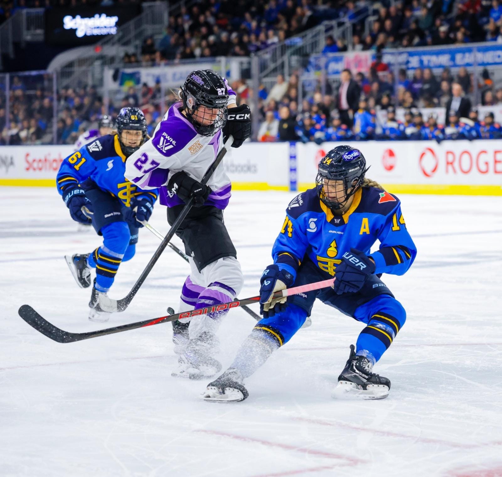 Fast (right in blue) looks at the puck as she stops while trying to knock it away from Heise (left in white). Heise is standing upright and dekeing around Fast, with the puck in her skates.