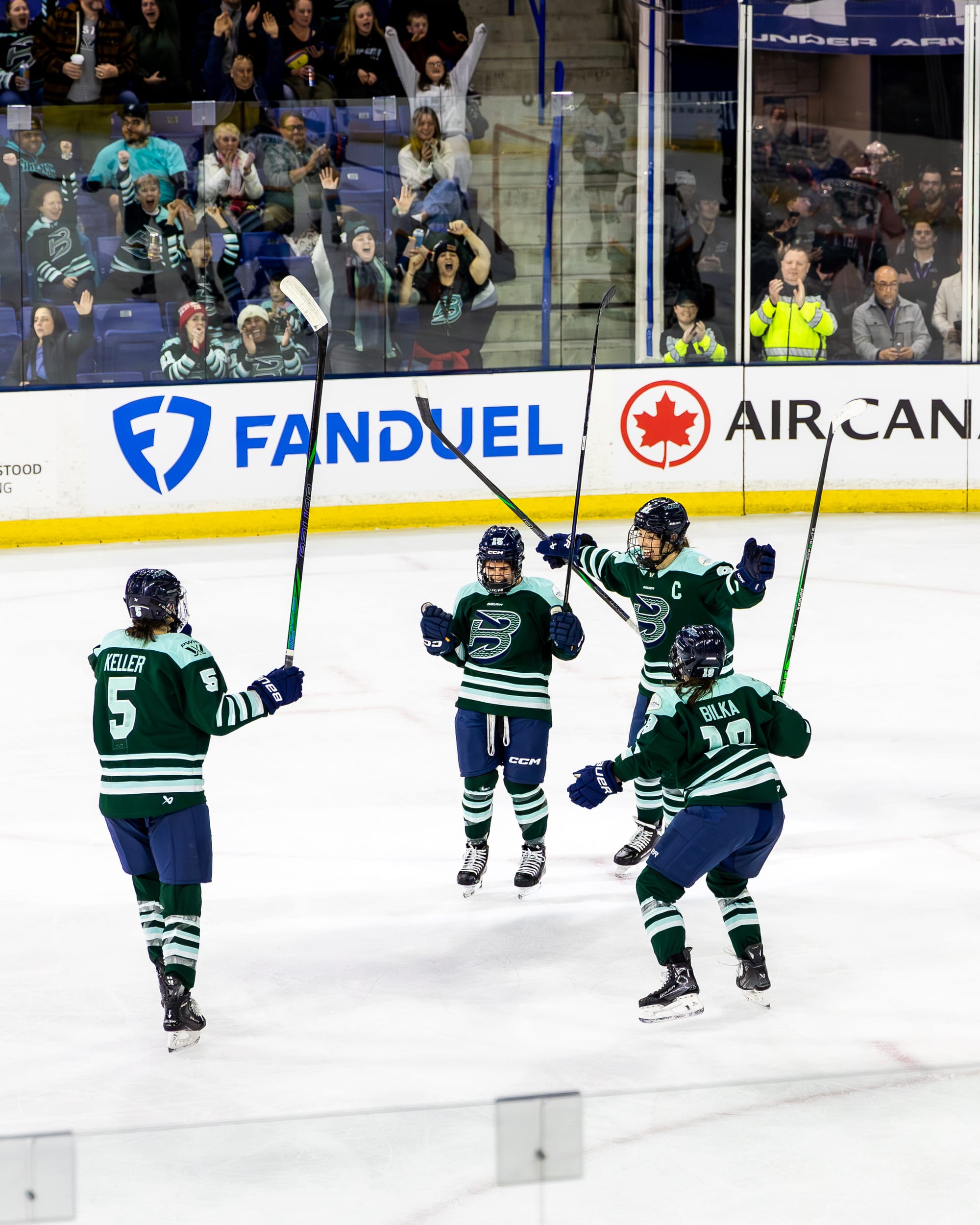 Bard (center) pumps her fists and yells in celebration. Knight and Bilka are coming in for a hug from the right, while Keller skates in from the left. They are all wearing green home uniforms.