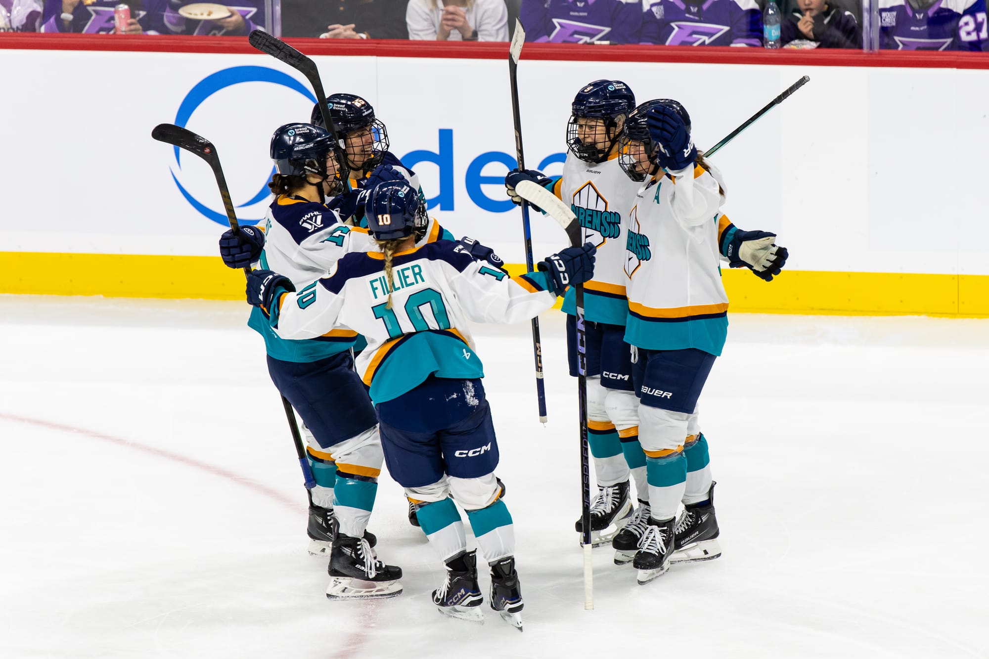 Five Sirens players celebrate a goal with a group hug. Four are side-to and their names/numbers are obscured, while Sarah Fillie is back-to. They are all wearing white away jerseys.