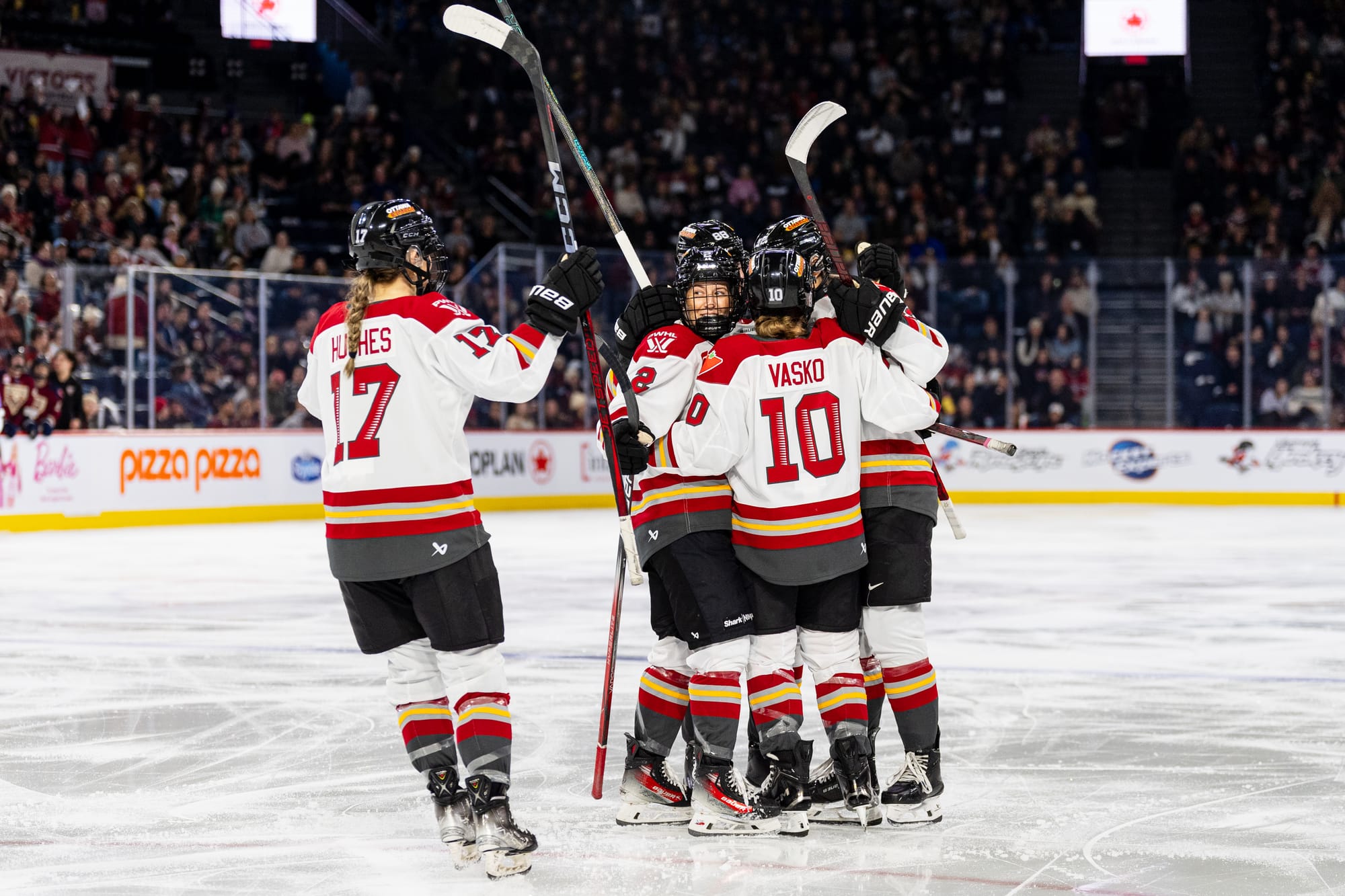 Five Charge players celebrate a goal with a group hug. Four are already in a tight hug, while Gabbie Hughes is skating into it. They are wearing white away jerseys.