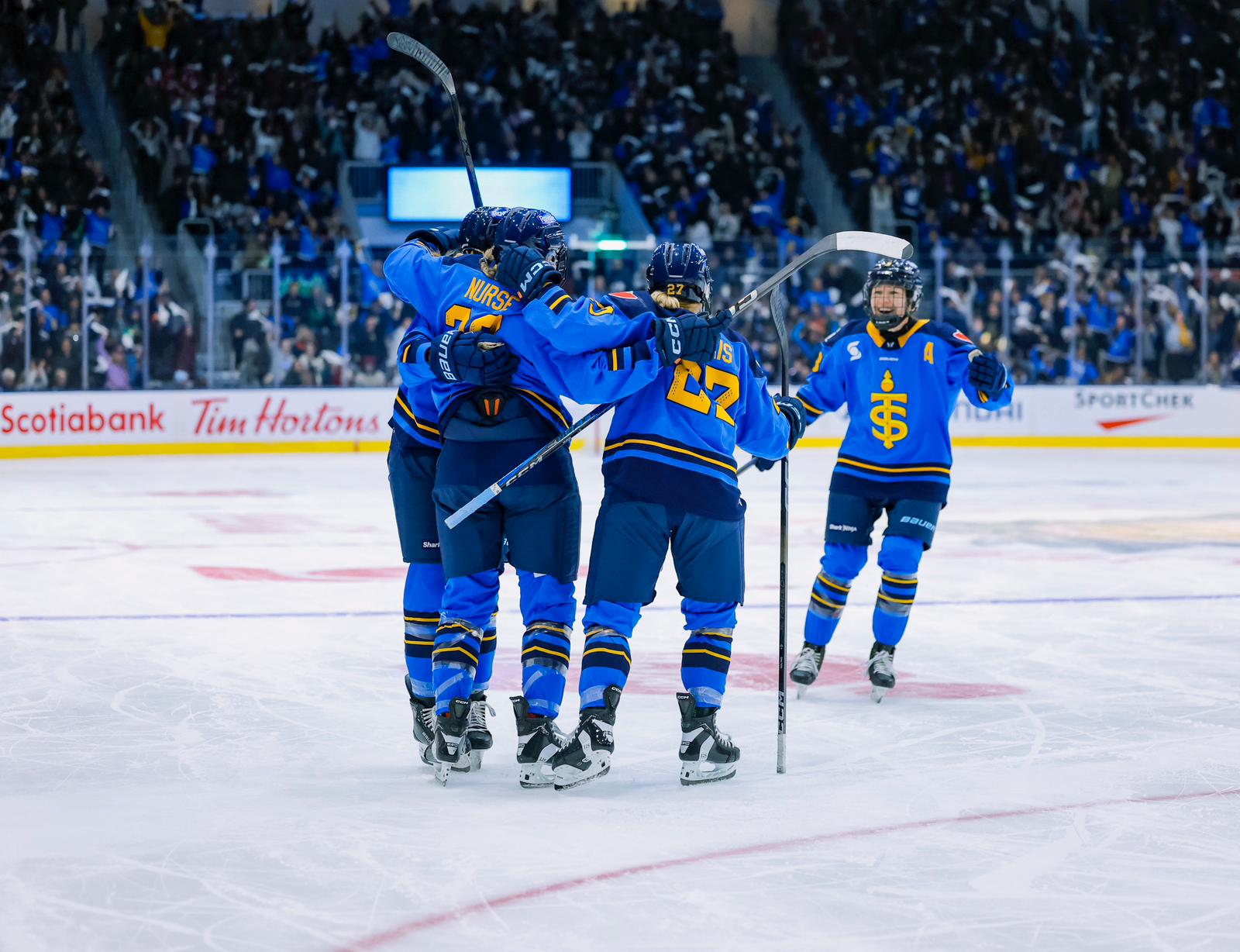 Four Sceptres players celebrate a goal with a group hug. Three are back-to, while one is just skating in. They are wearing blue home jerseys. 