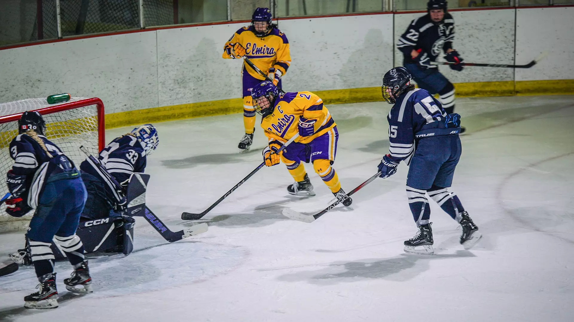 Two Elmira hockey players, in gold jerseys and purple pants, are near the Southern Maine goal. The goal is defended by three players and a goalie, all dressed in navy.