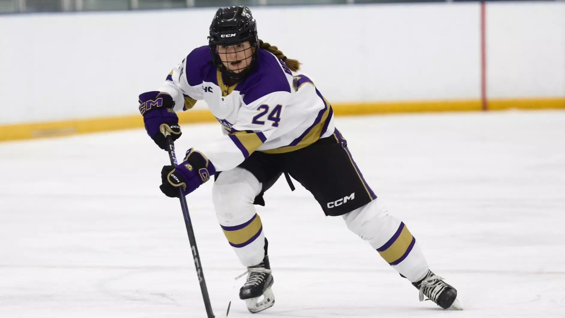 The hockey player is left-handed. She wears a white jersey, #24, with purple epaulets and gold trim. Her pants, and gloves are black with purple and gold trim. Her socks are white, with wide gold stripes with narrow purple borders. Her sleeves and waist have similar stripes. Her medium-brown hair is braided and hangs down below her black helmet.