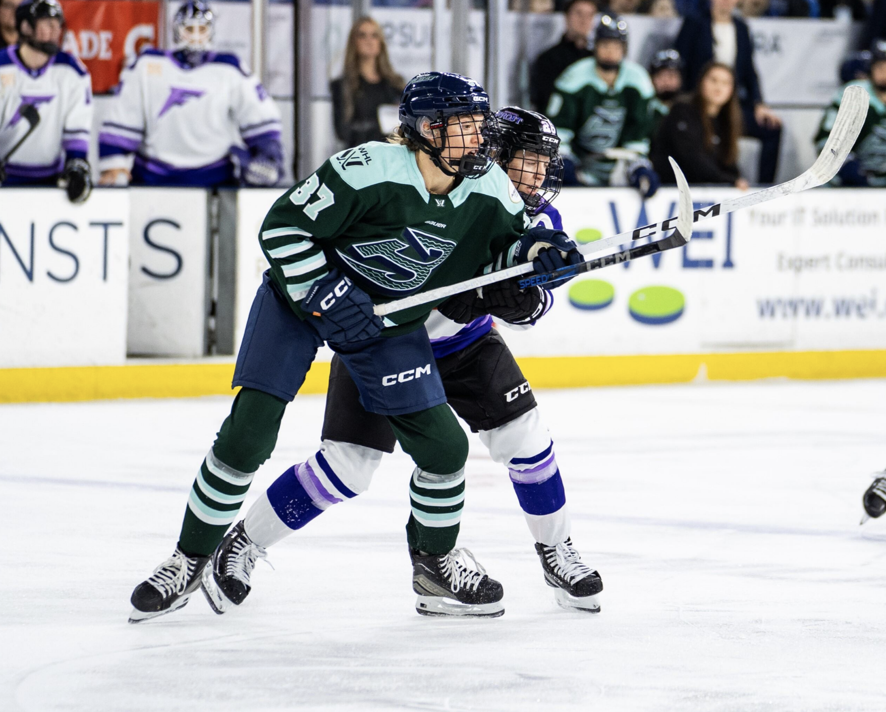 Shirley (front in green) and Coyne Schofield (behind in white) battle for positioning near the blue line. They are both watching the play, which is out of frame.