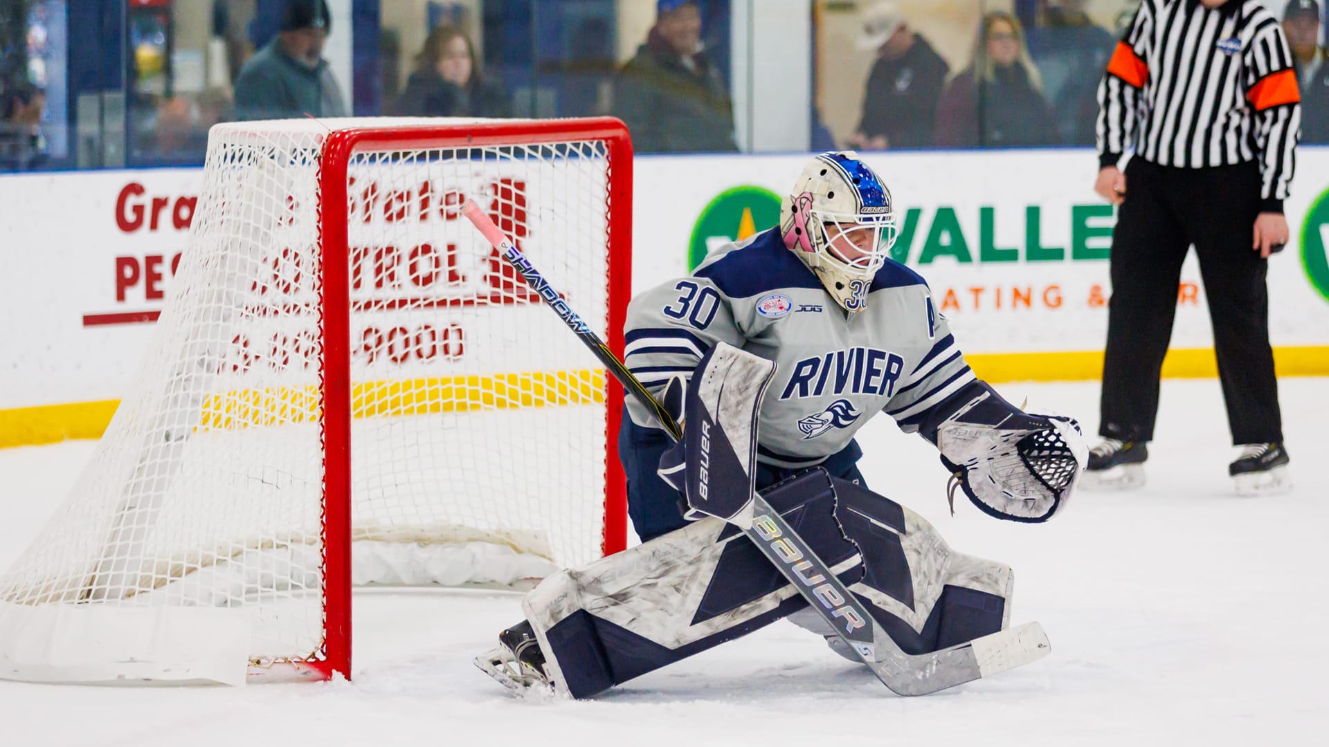 Goalie #30 is about to go down in the butterfly position in front of her own net. She's wearing a gray "away" jersey with navy trim. Her pads are white and navy. Her helmet is white with a royal blue racing stripe down the top of the head and a pink breast cancer awareness ribbon on the side. Her stick is ready to cover her five-hole, with the blade halfway covered in white tape and the top of the handle taped in pink. 