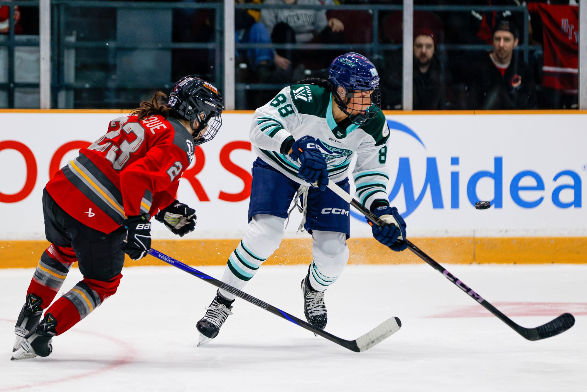 Adzija  (right in white) skates against Larocque (left in red). Adzija is carrying the puck to her left and looking at it, while Larcoque is skating in to poke it away.