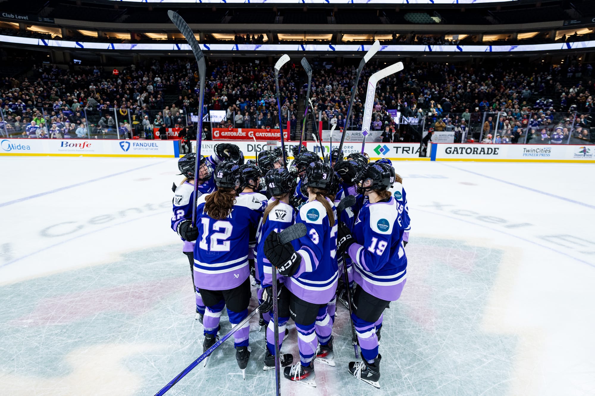 The whole Frost team celebrates a win with a tight huddle at center ice. Many are raising their sticks above their heads. They are all wearing purple home uniforms.