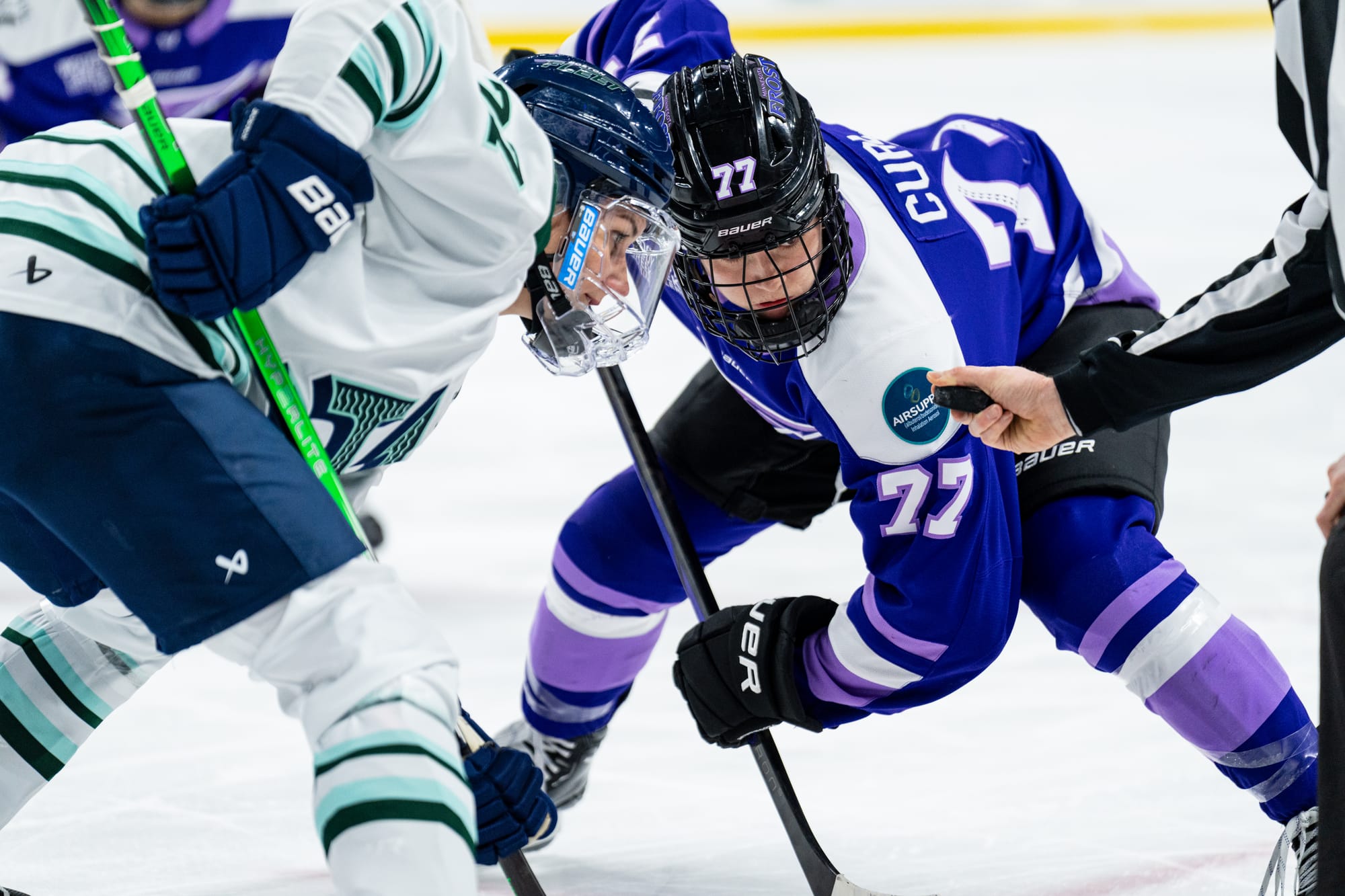 Tapani (left in white) and Curl-Salemme (right in purple) look at the puck. They're crouched with their sticks down on the ice, positioned in the faceoff dot out of frame.  