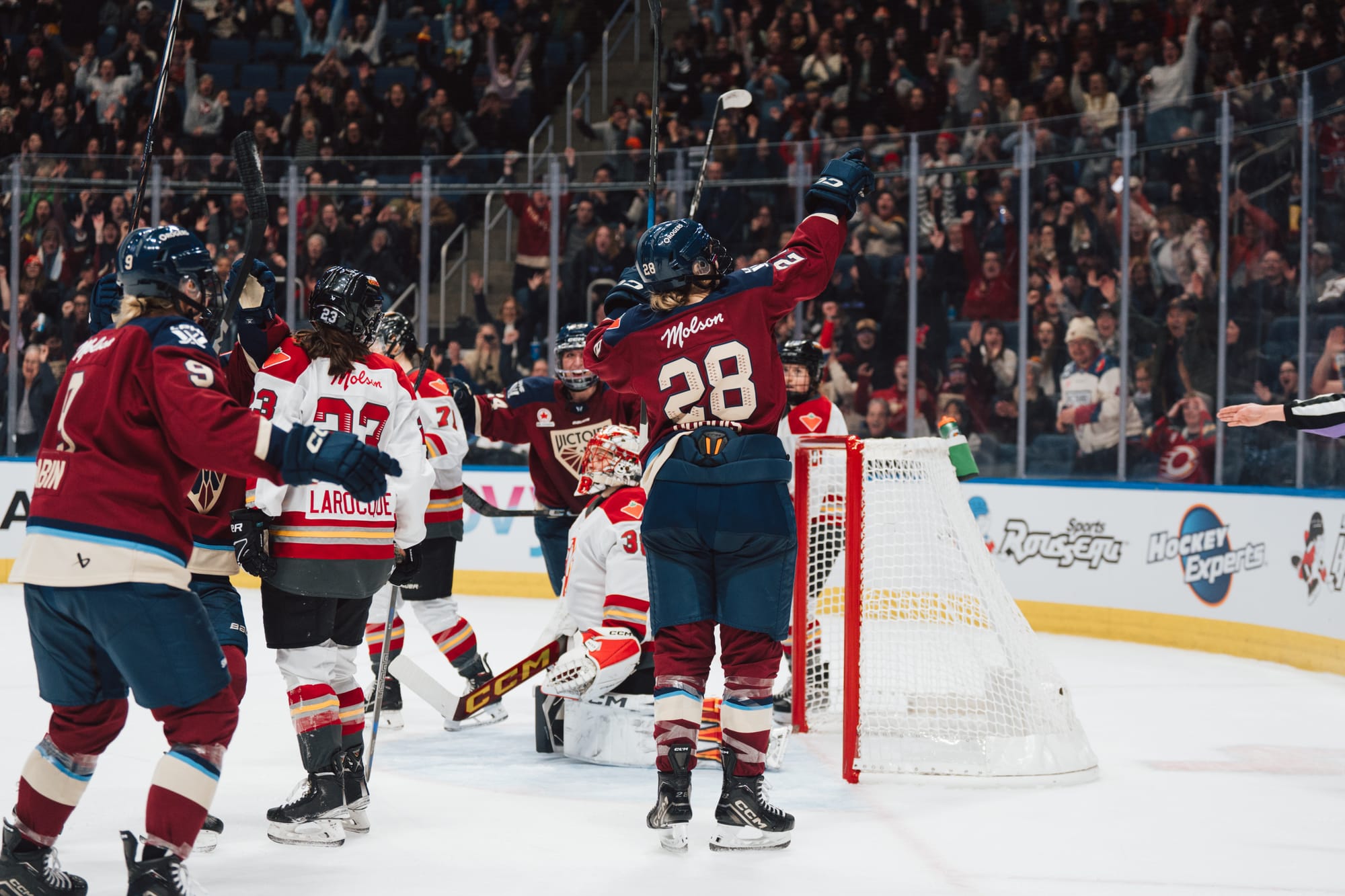 Dubois points up into the crowd. Her back is to the camera and she is facing toward the end boards. She is wearing a maroon home jersey, and there are two other Montréal players skating toward her.