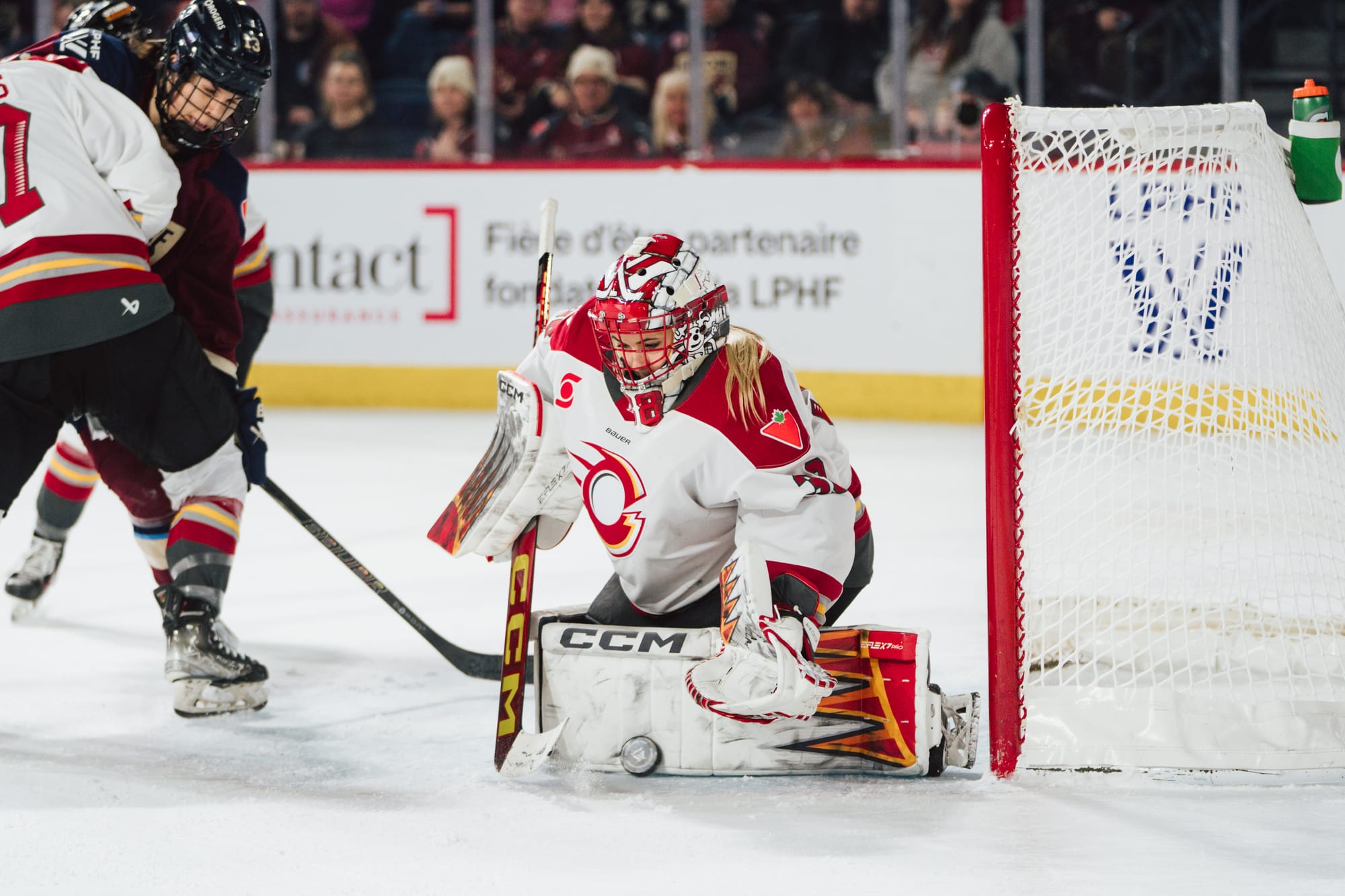 Maschmeyer makes a save with her left pad from her knees. She is moving to cover the puck and wearing a white away uniform.