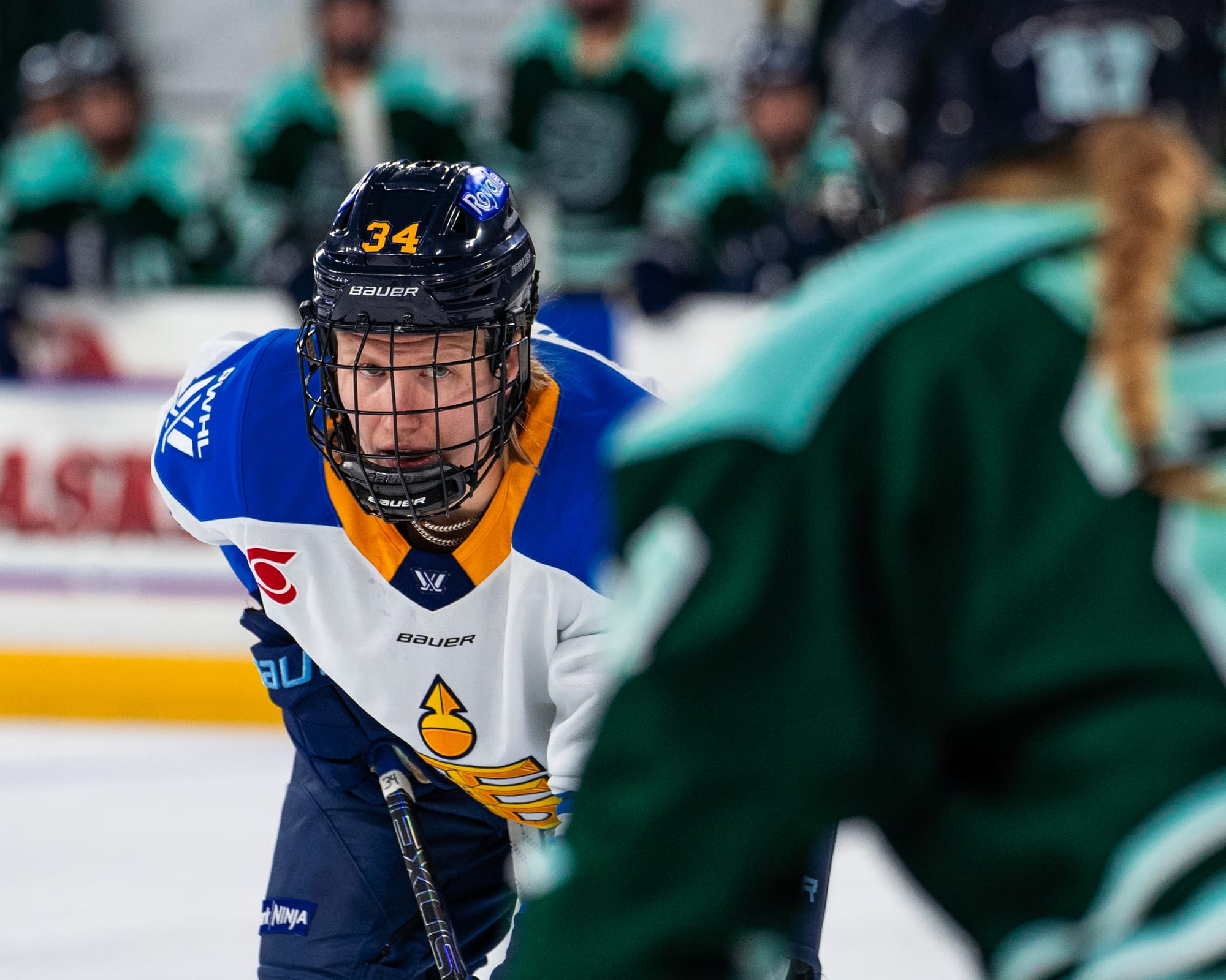 Miller looks forward from her wing spot on the hashmarks of a circle. She is leaning on her stick prepared for the puck to drop and wearing a white away uniform.