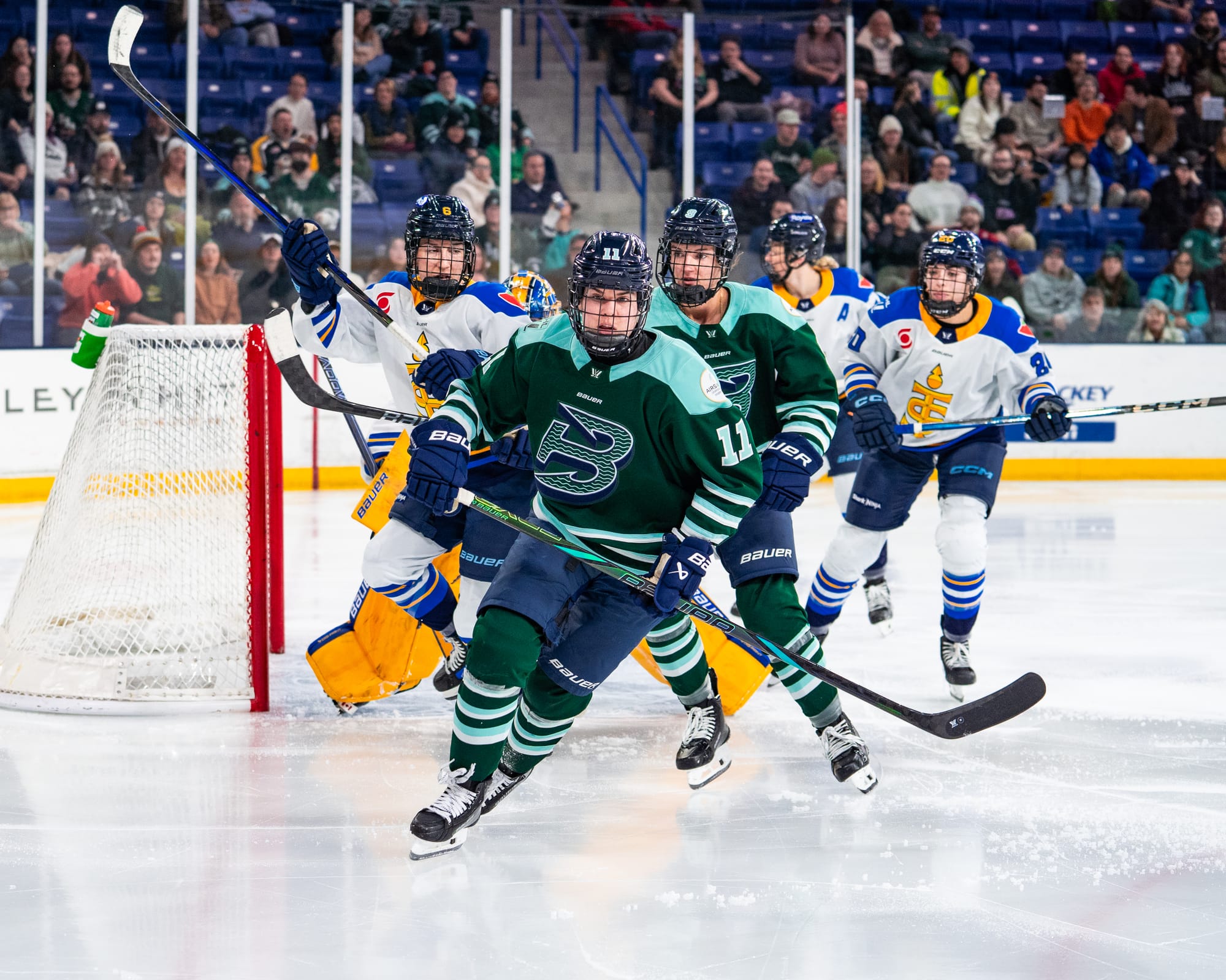 Müller is facing the camera and skating towards the corner, likely to battle for the puck. She is wearing a green home uniform, and there is another Fleet player along with three Sceptres players trailing her.