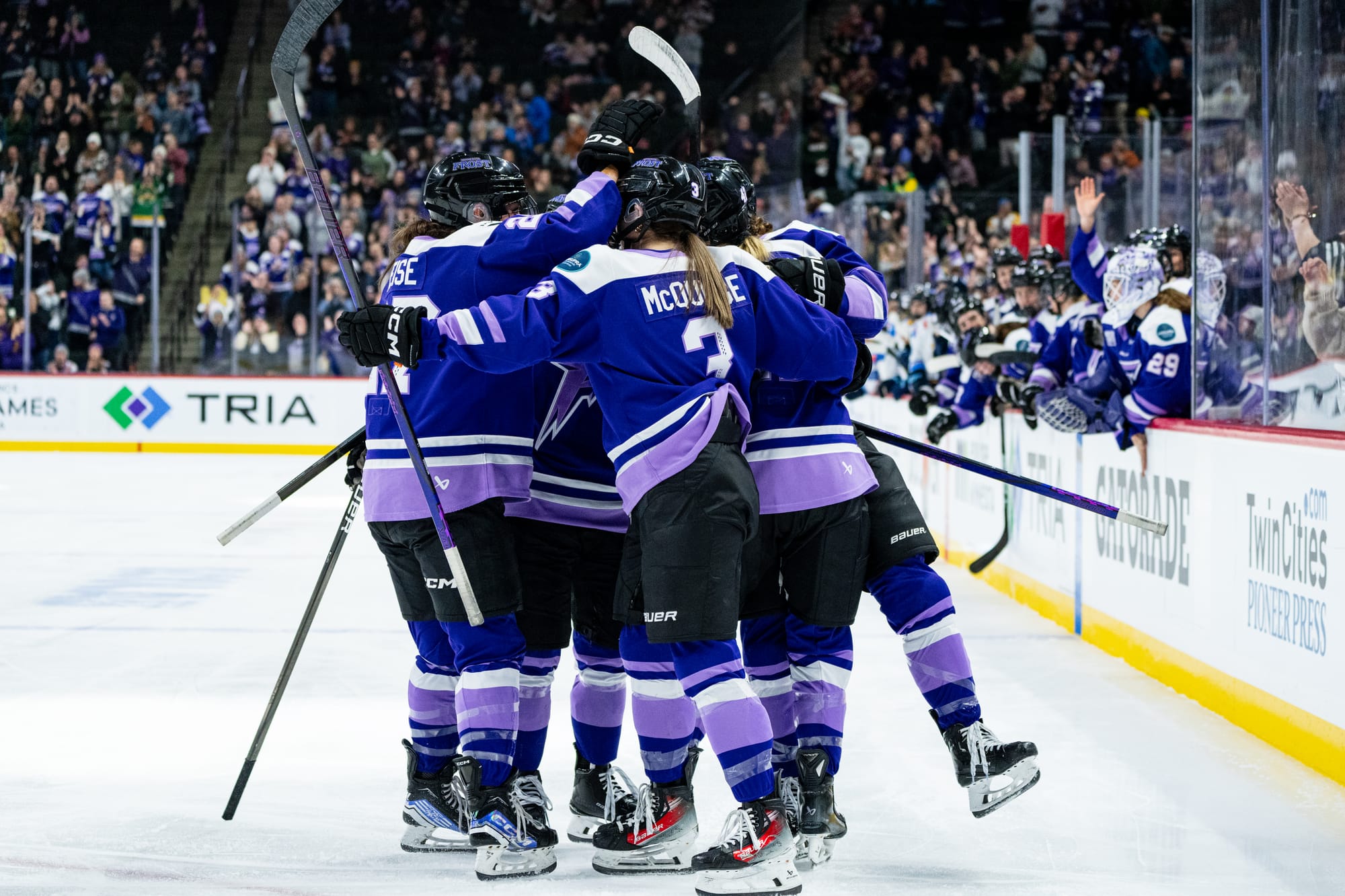 Frost skaters celebrate a goal with a tight group hug at the top of one of the circles. They are wearing purple home uniforms. 