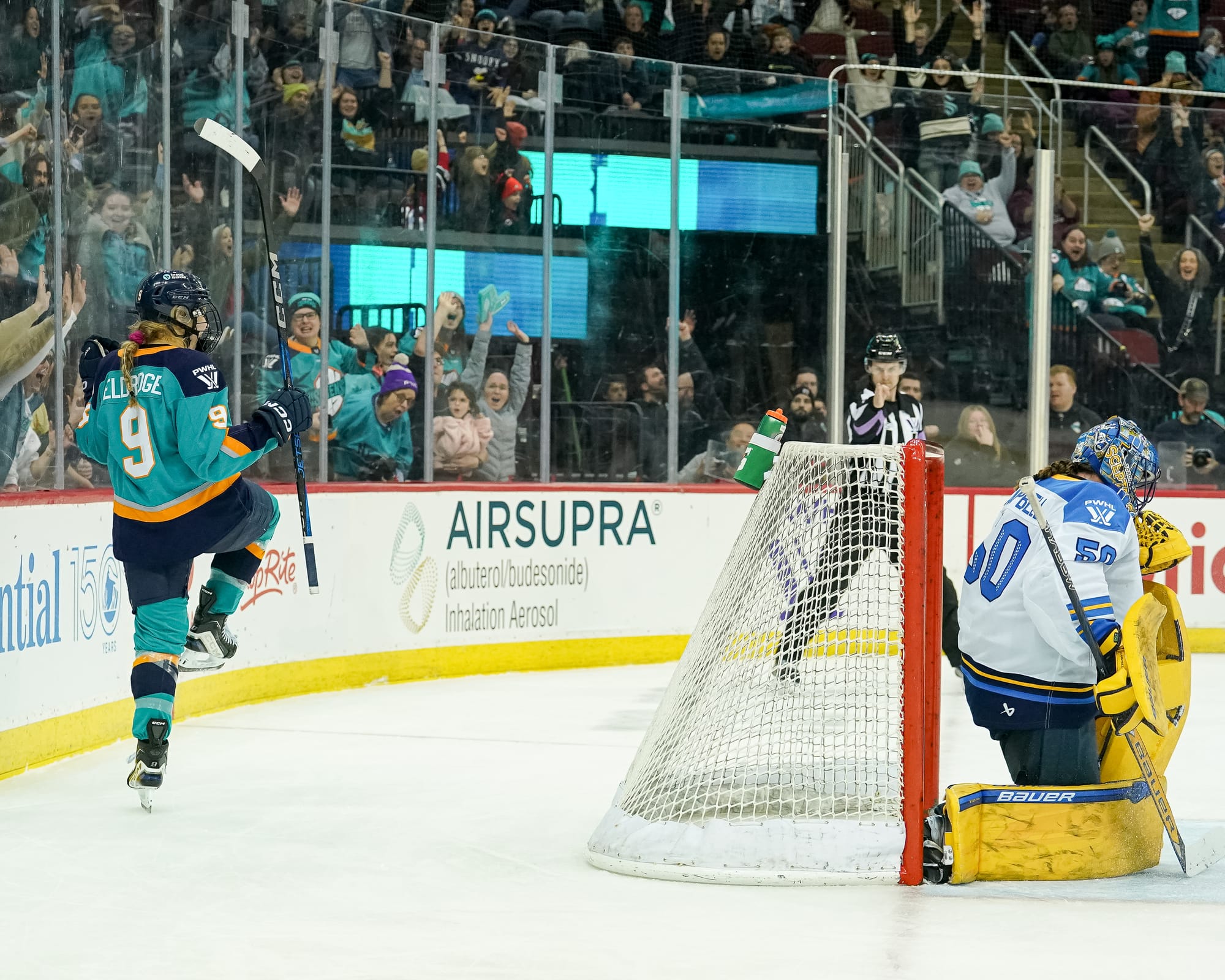 Eldridge lifts her right leg, raises her stick, and pumps her fists in celebration behind the goal. She is wearing a teal home uniform.