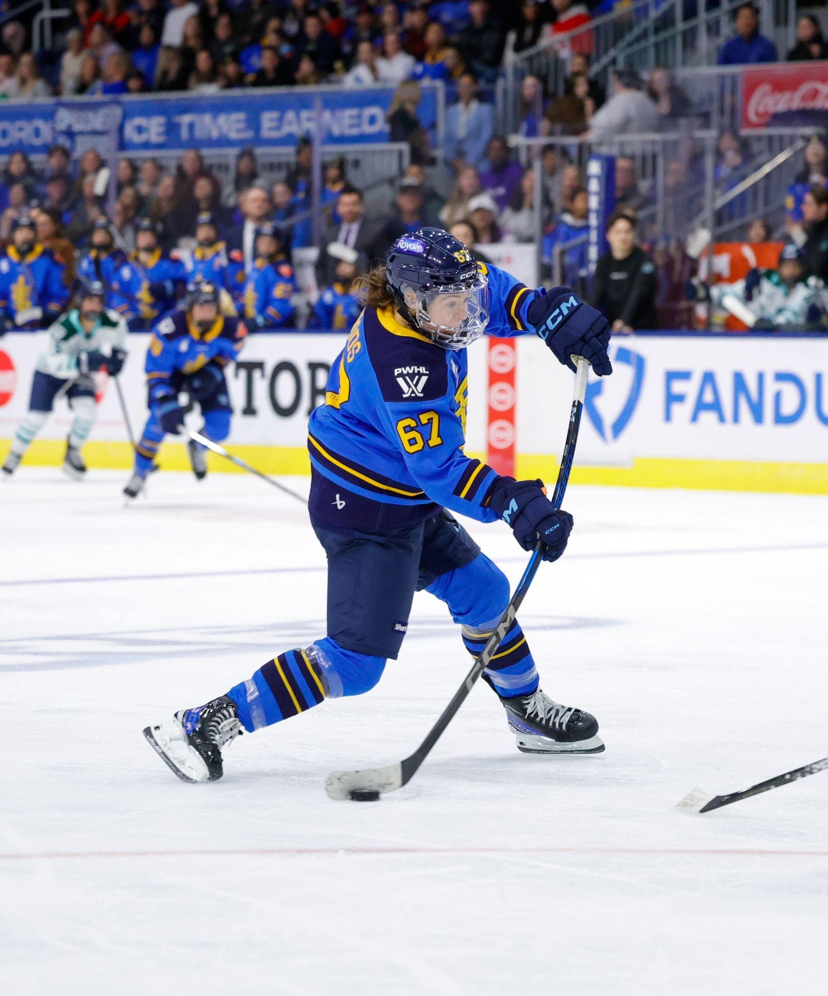 Woods flexes her stick as she shoots the puck. Her knees are bent and she's transferring her weight to her front (left) leg. She is wearing a blue home uniform.