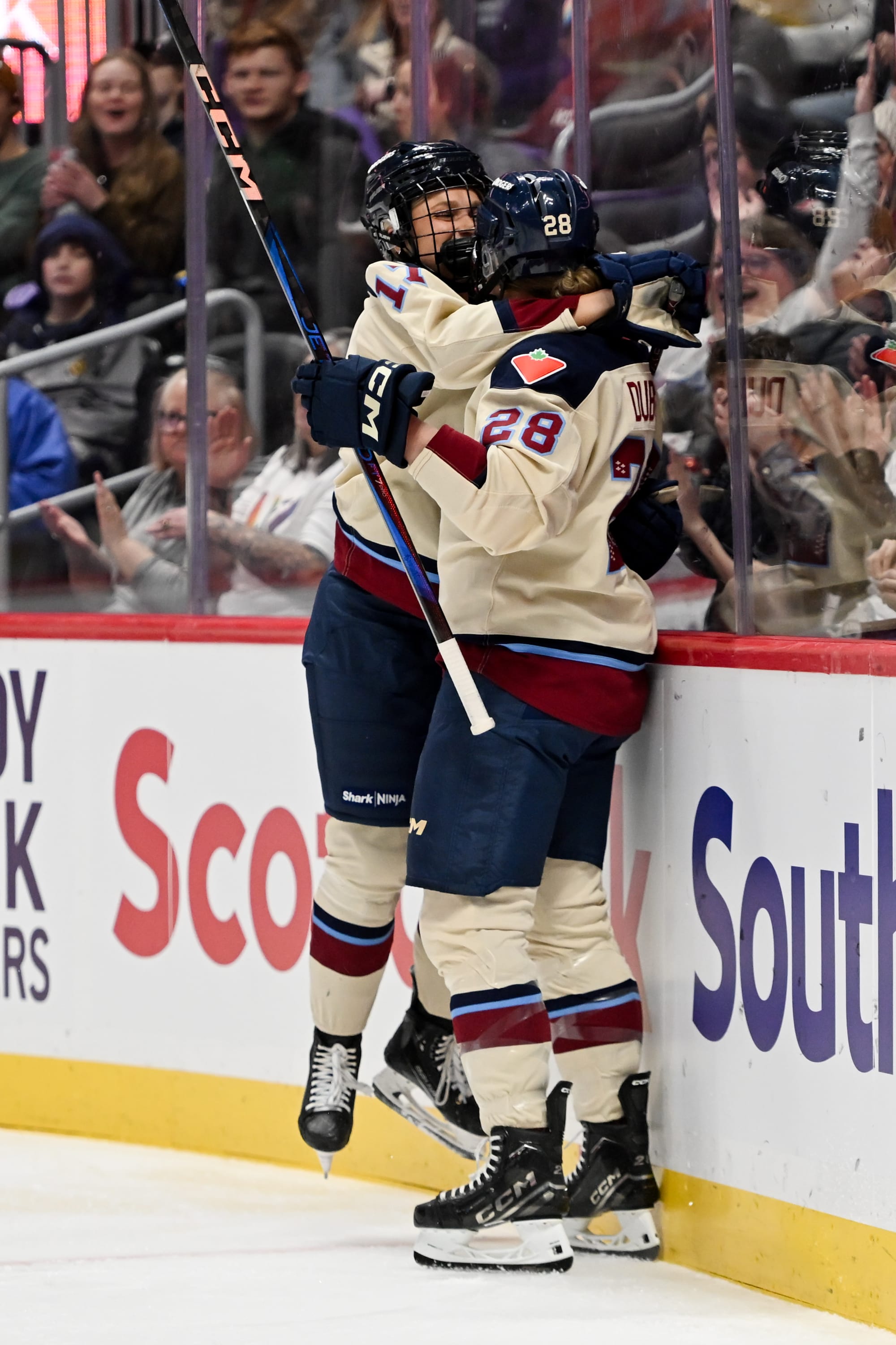 Two Montréal players celebrate a goal with a hug along the end boards. One is midair as they jump into it. They are wearing cream away uniforms.