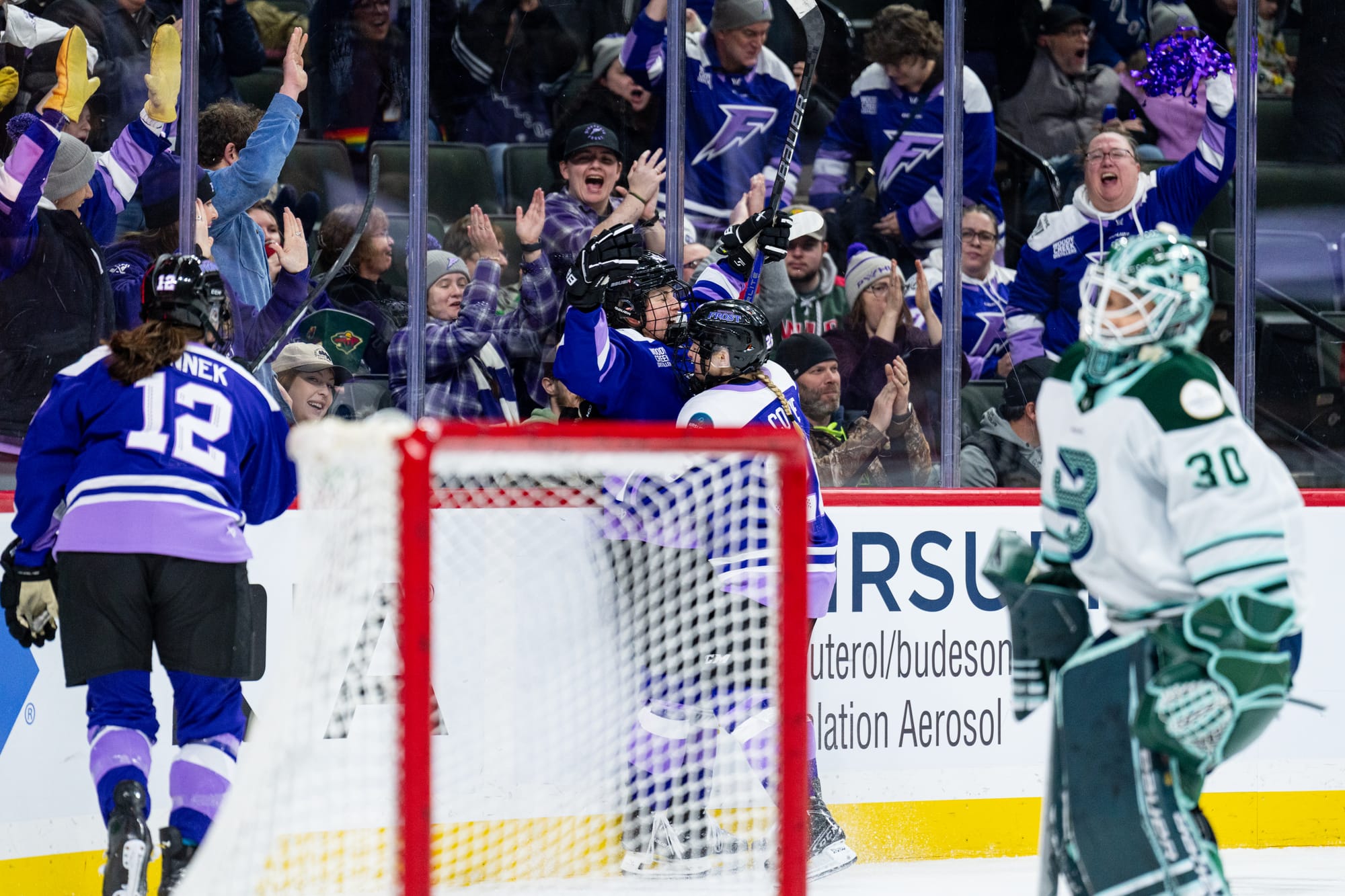 Two Minnesota players celebrate with a group hug behind the goal. Another is skating in to join. They are wearing purple home uniforms.