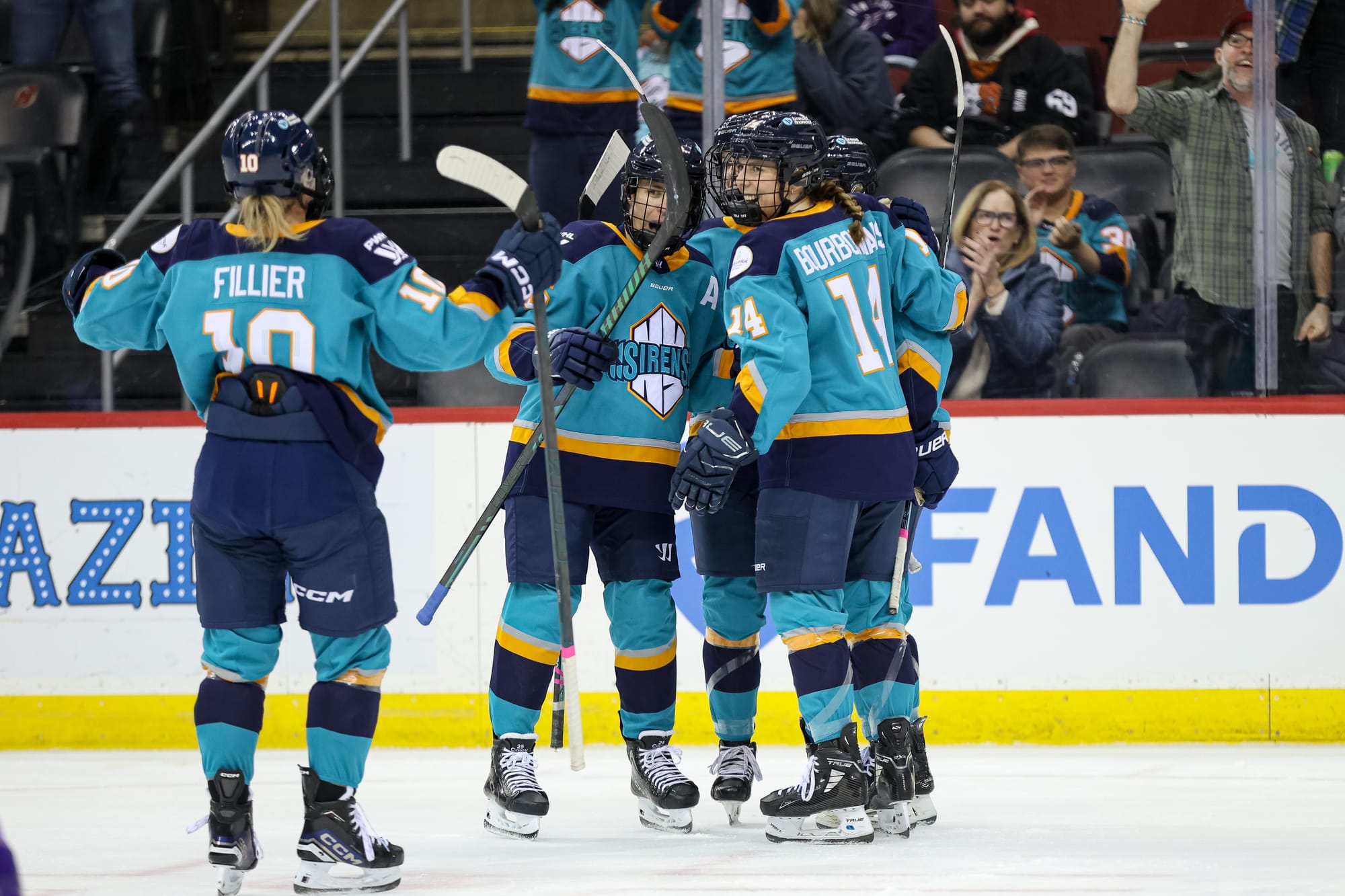 Sirens players celebrate a goal with a group hug. Four are already part of it, while Fillier is skating in from the left. They are wearing teal home uniforms.